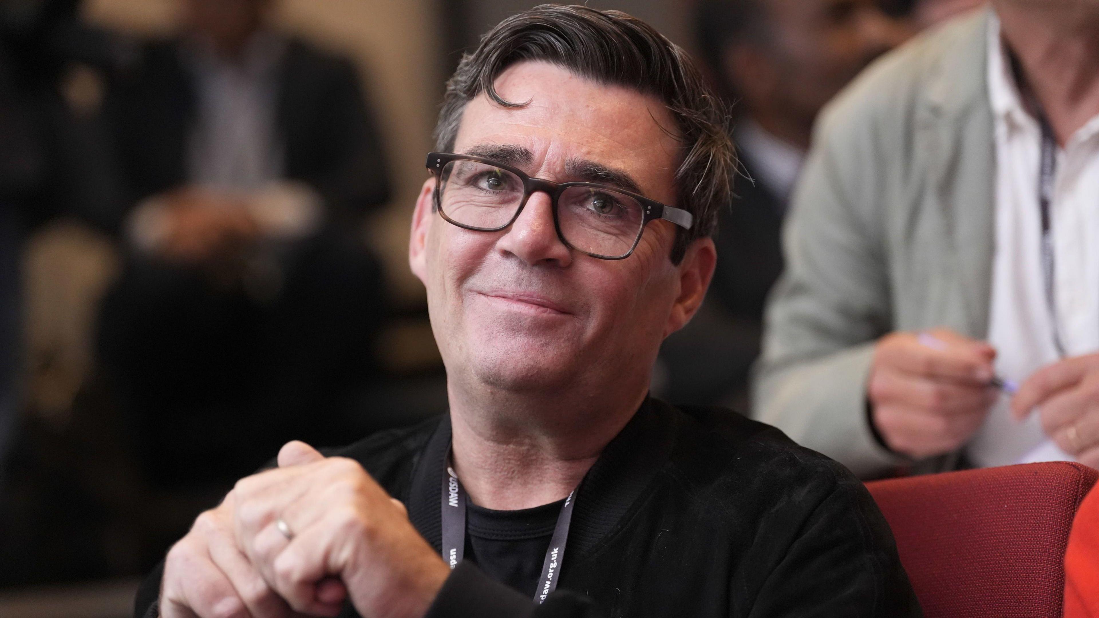 A smiling Andy Burnham looking off into the distance. He is wearing dark rimmed glasses and has dark hair combed in a side shed.