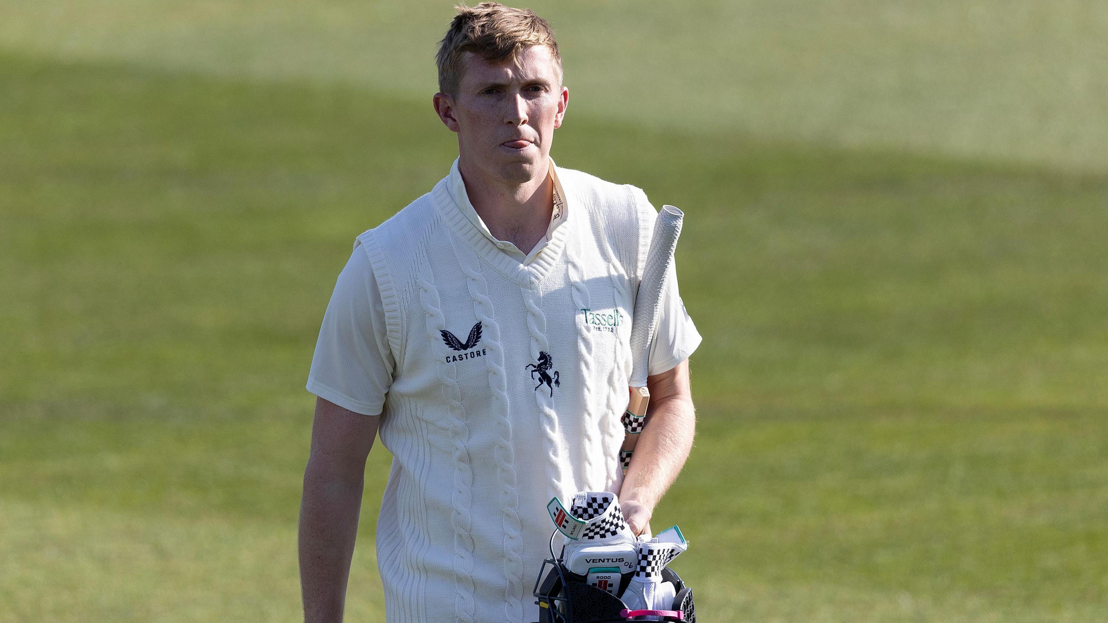 Zak Crawley walks off the cricket ground, dressed in a white short-sleeved top and white knitted vest, carrying a blue helmet and white and black gloves