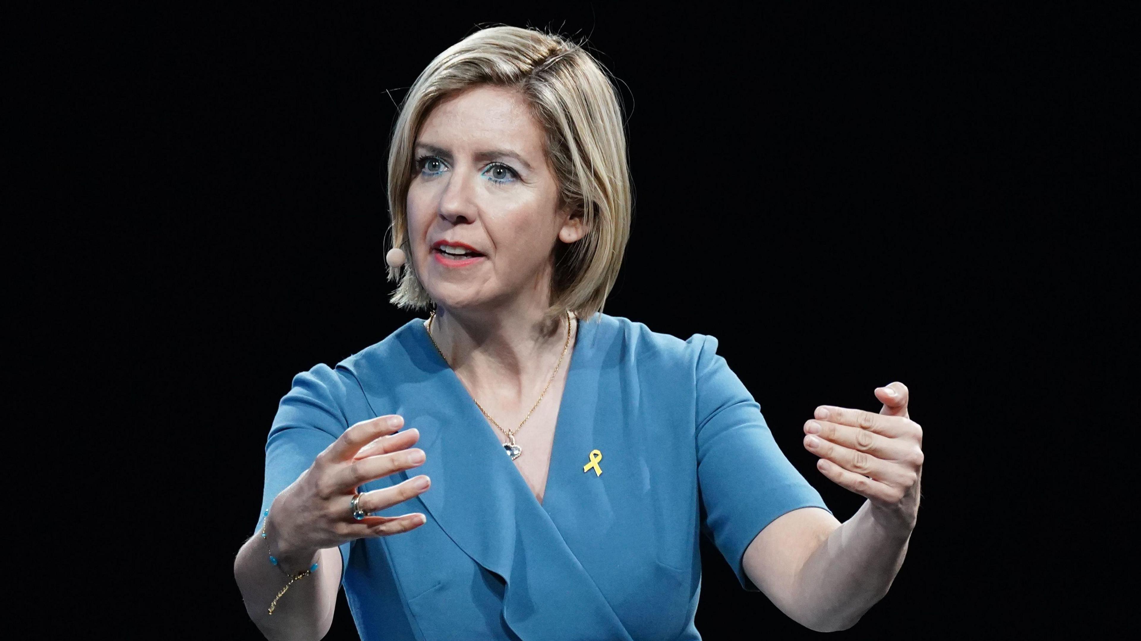 A woman with bobbed blonde hair delivers a speech while wearing a wireless microphone. She has her arms outstretched in a gesture and wears a blue dress.