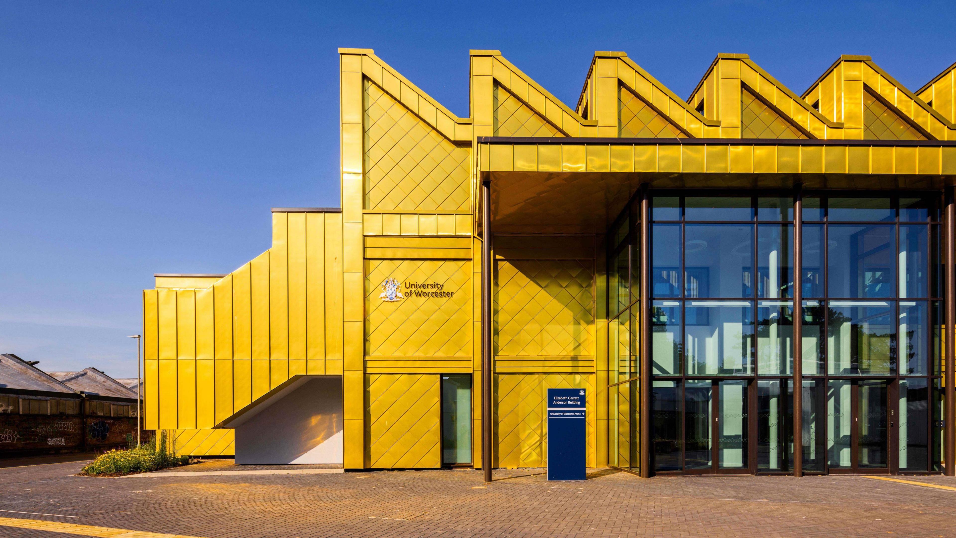 A yellow building with a glass entrance on the right. The building has a triangular feature that runs along the top of the roof. The sky is blue in the background.