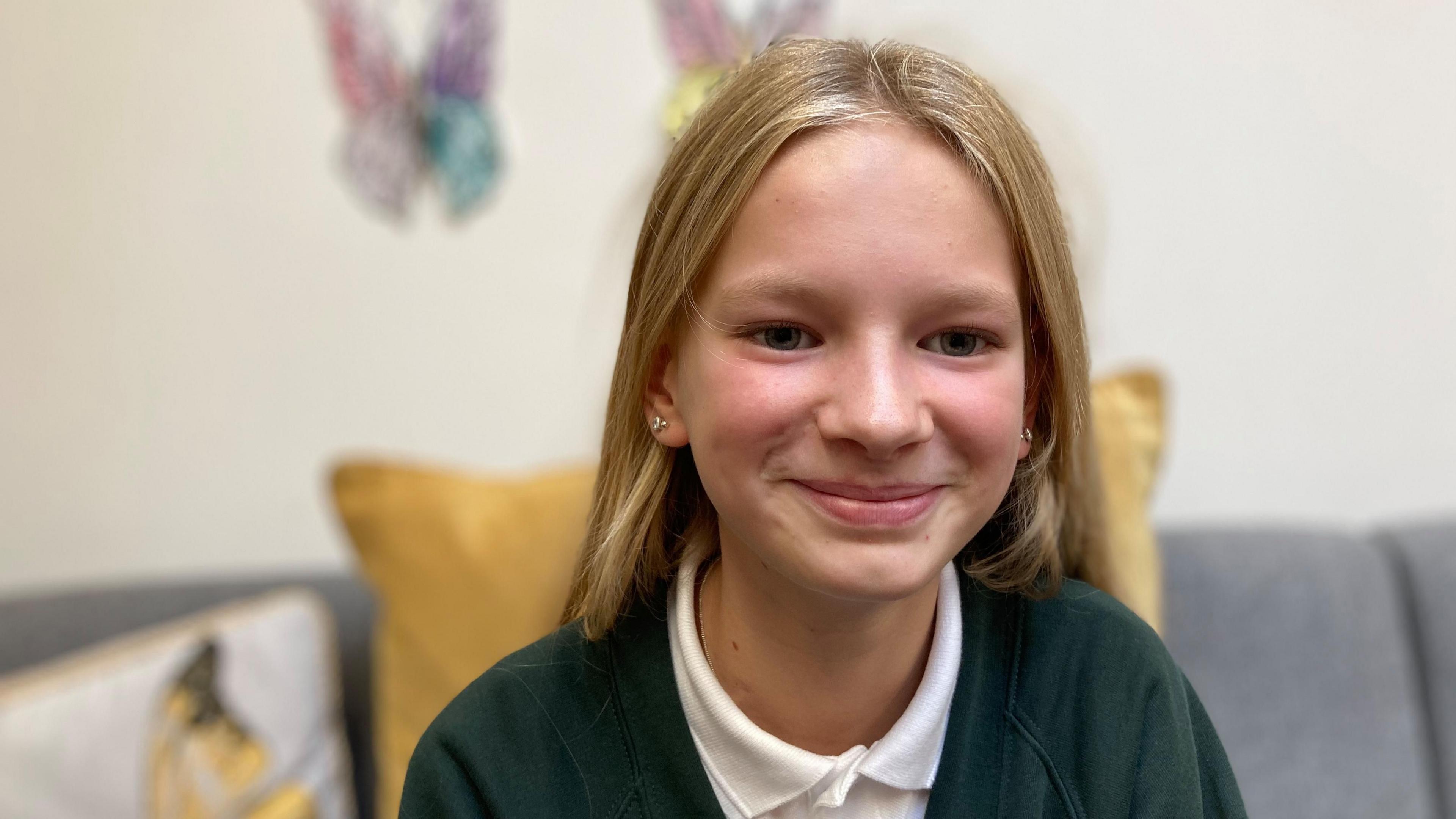 Maggie smiles at the camera as she is photographed in her school uniform. She has blonde hair and is sitting on a sofa.