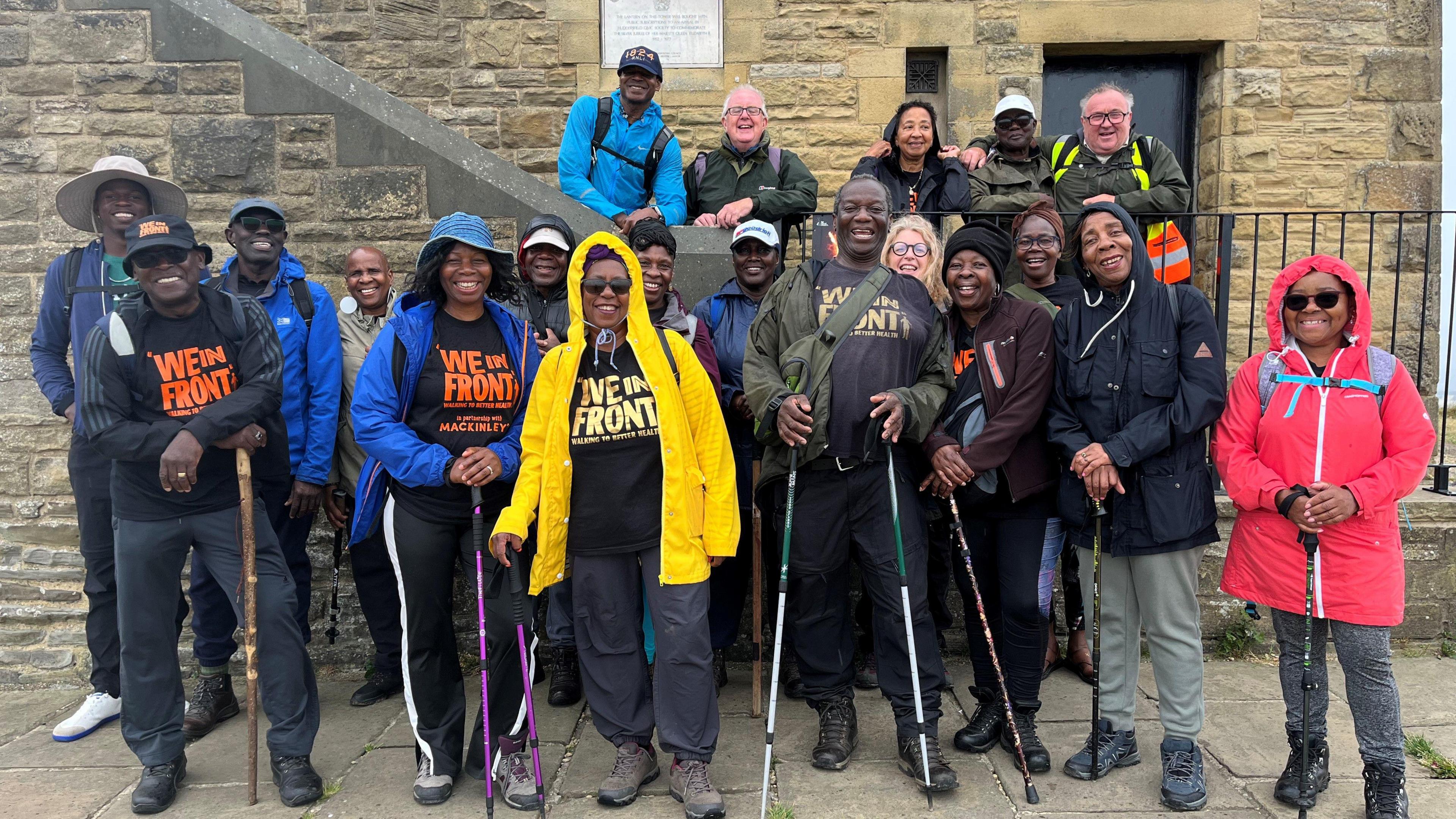 A group of twenty people, clearly walkers (many with walking poles, all wearing walking boots and raincoats) are gathered together. Several have T-shirts with the groups name 'We In Front' on them. They are stood in front of Castle Hill in Huddersfield, and are all looking directly at the camera smiling. They are aged between 50 and 80, with one younger member.