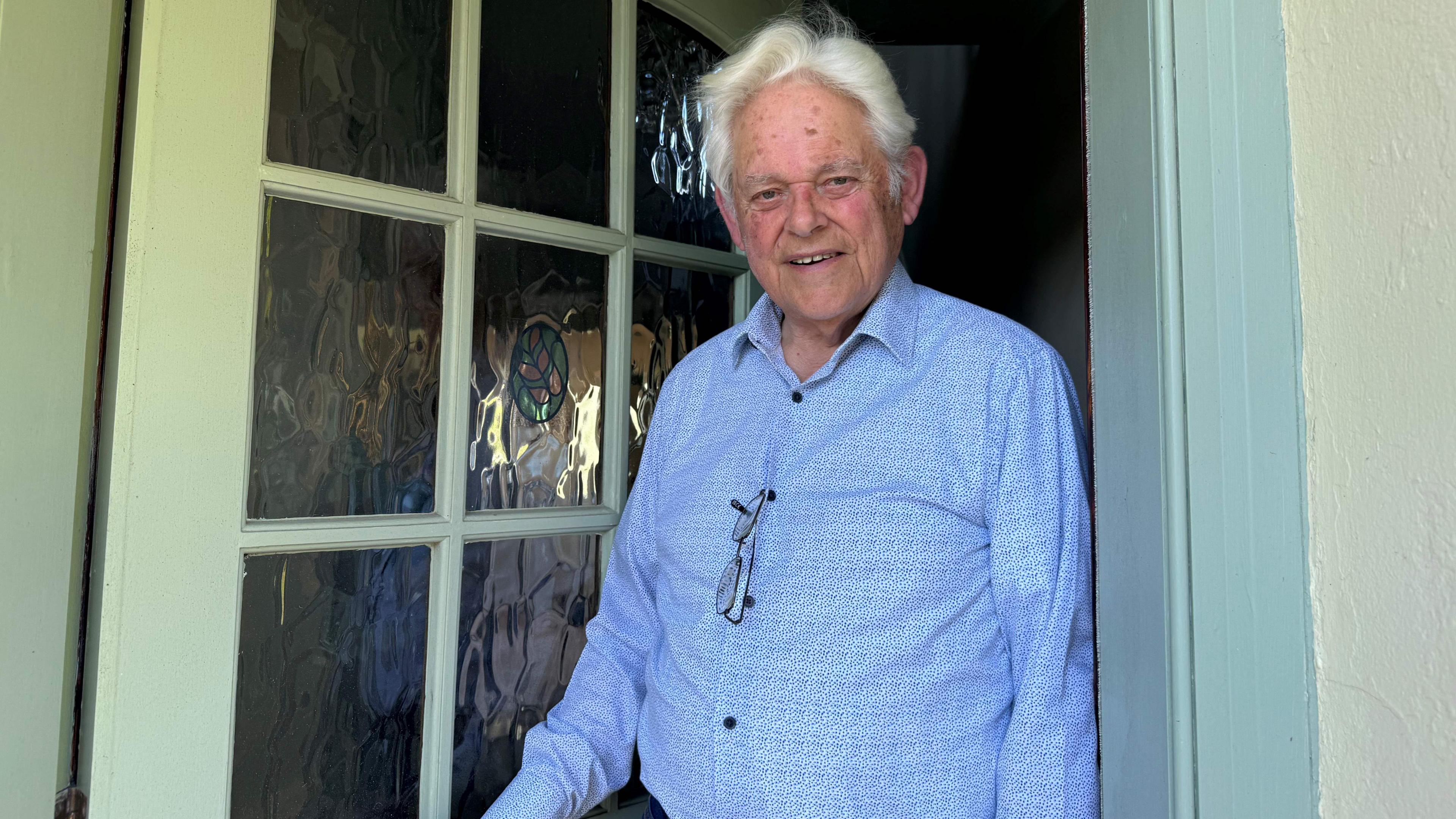 Pierre Tostevin standing in his doorway, he is wearing a blue shirt