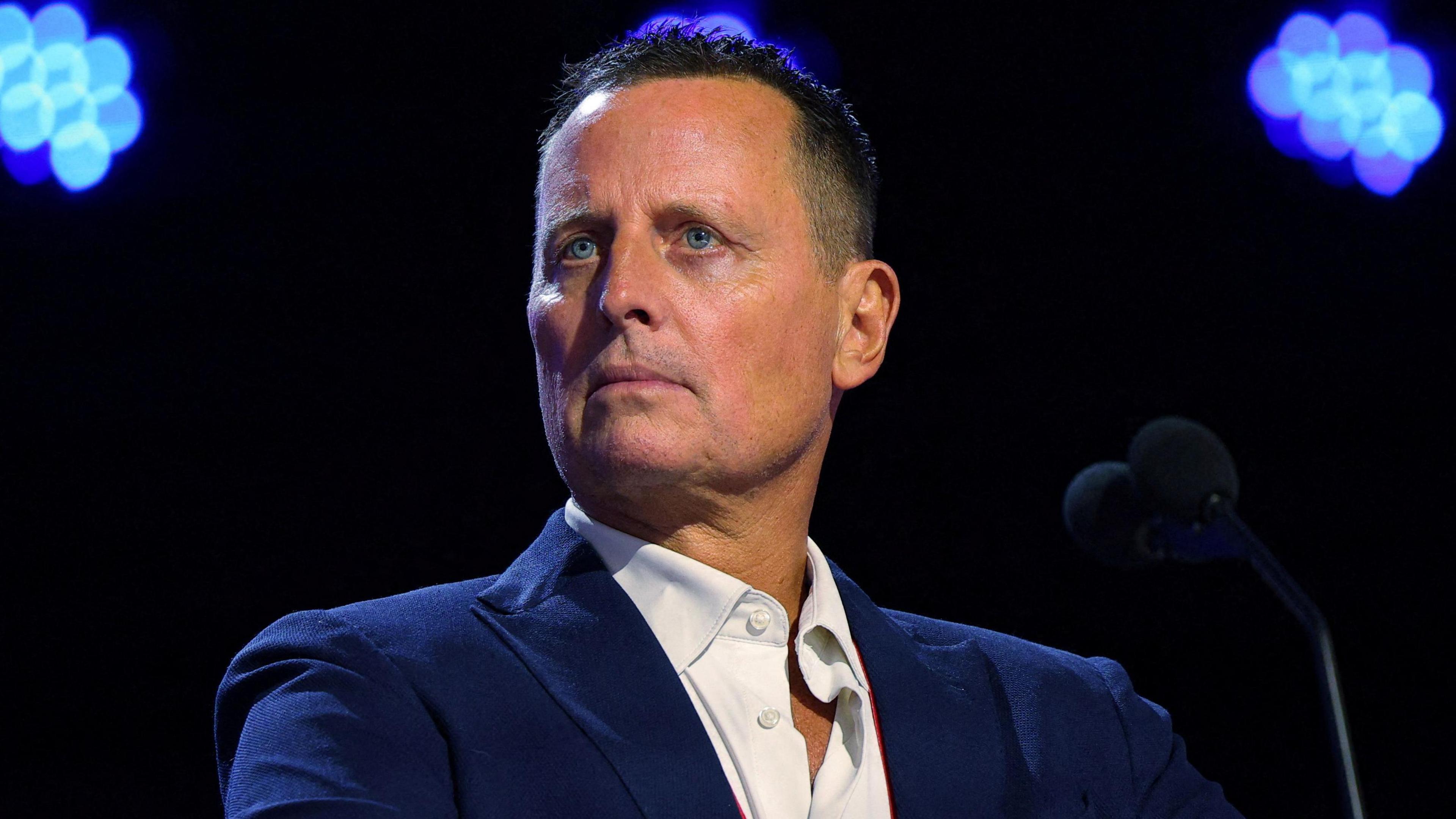 Richard Grenell, who has short brown hair, wears a blue suit jacket and white shirt, as she stands in front of a microphone onstage at a convention