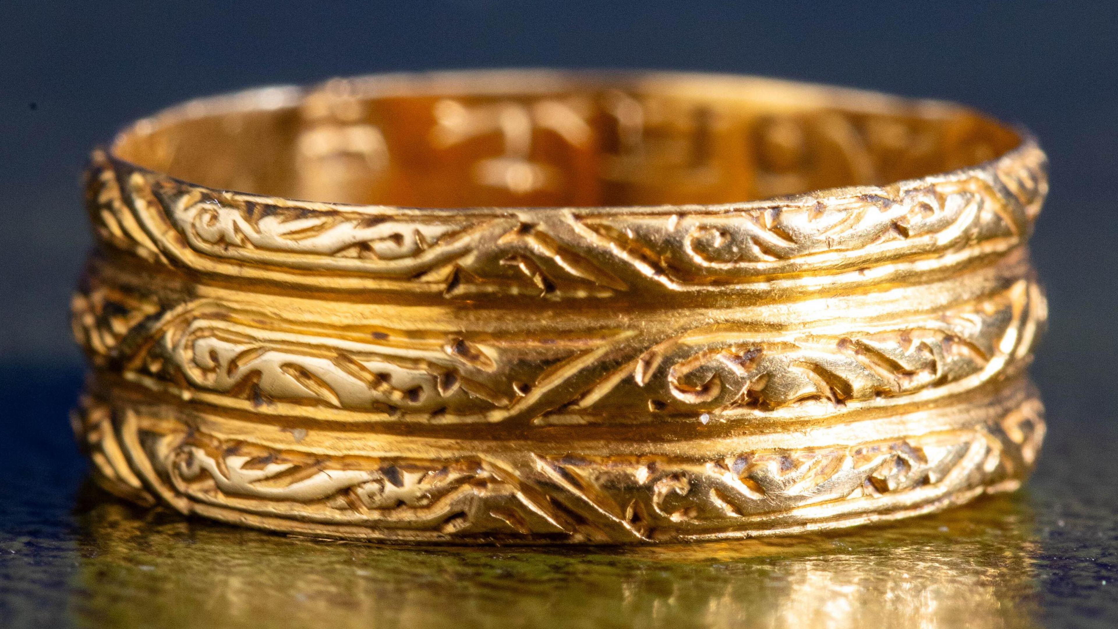 Three-banded gold ring, showing scrolling foliage on all three bands. The ring is on a table in which the reflection is visible.