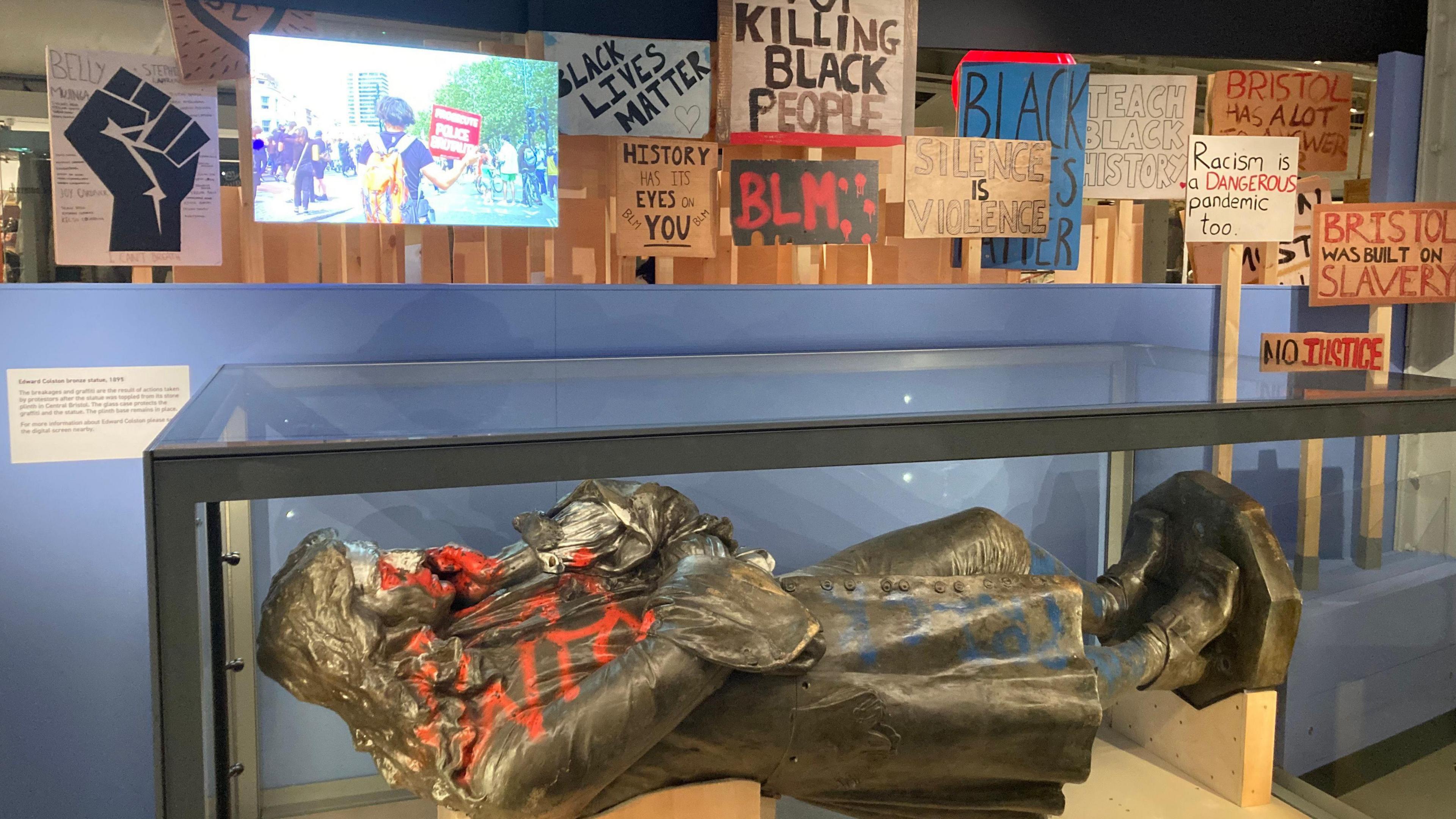 Metal statue of a man in a long coat with long hair. He has crossed arms and has is chin in one of his hands, and he appears to be looking down. The statue is on its side in a glass display tank in a museum, with placards from Black Lives Matter protests behind it.