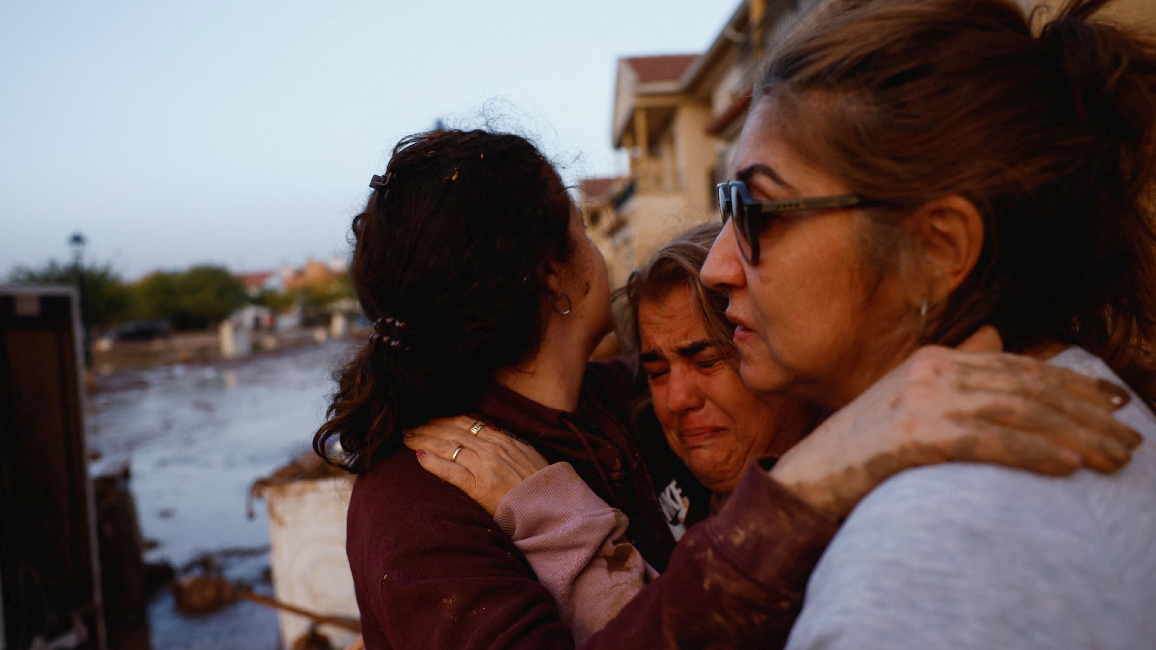 Three women embrace, with one crying, as they stand in front of a house and floodwater 