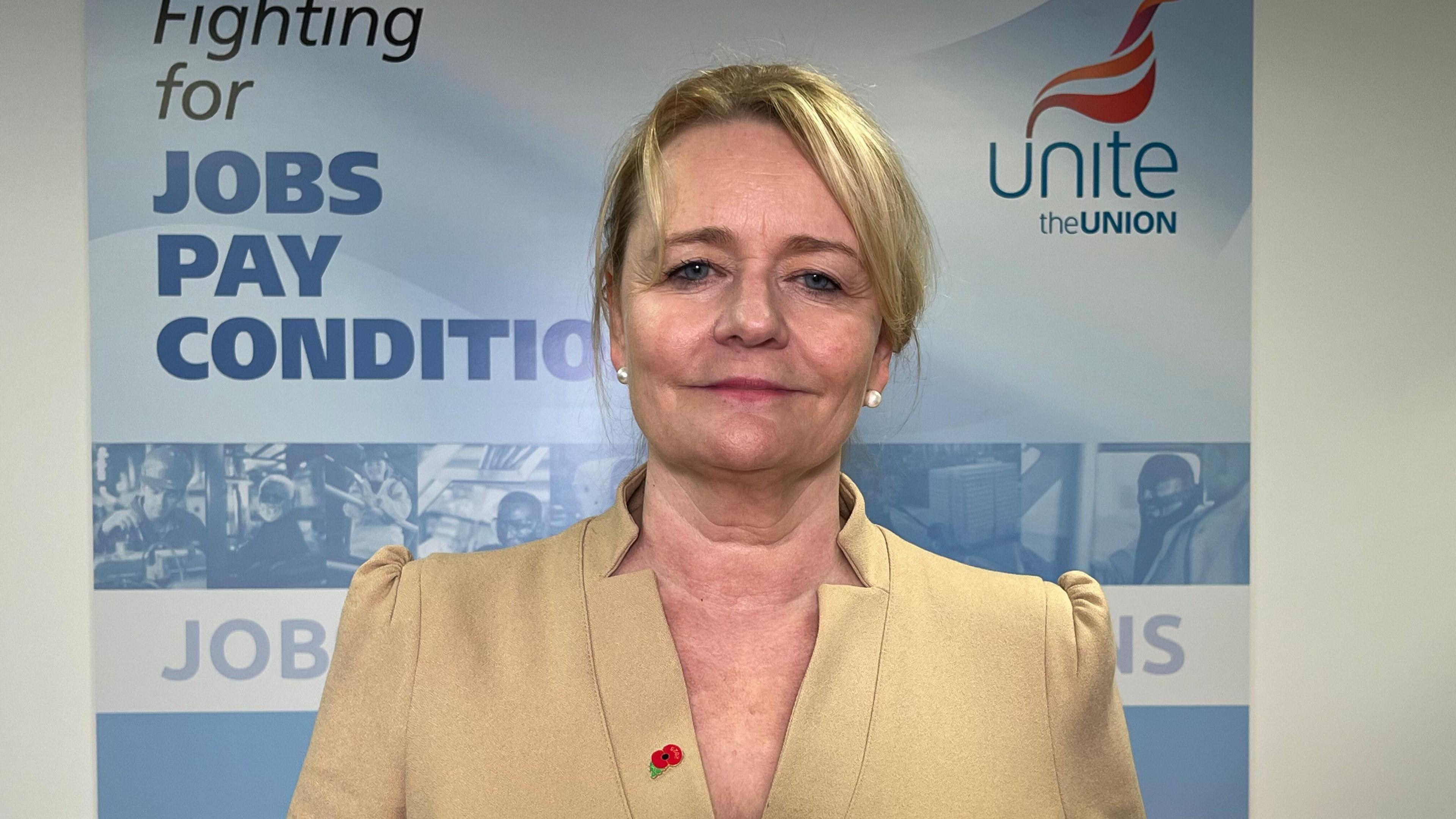 Sharon Graham standing in front of a banner with Unite's branding on it - she is wearing a cream / gold structured top,  has blond hair, is wearing pearl-like earrings, has a poppy on her lapel, and has a neutral expression