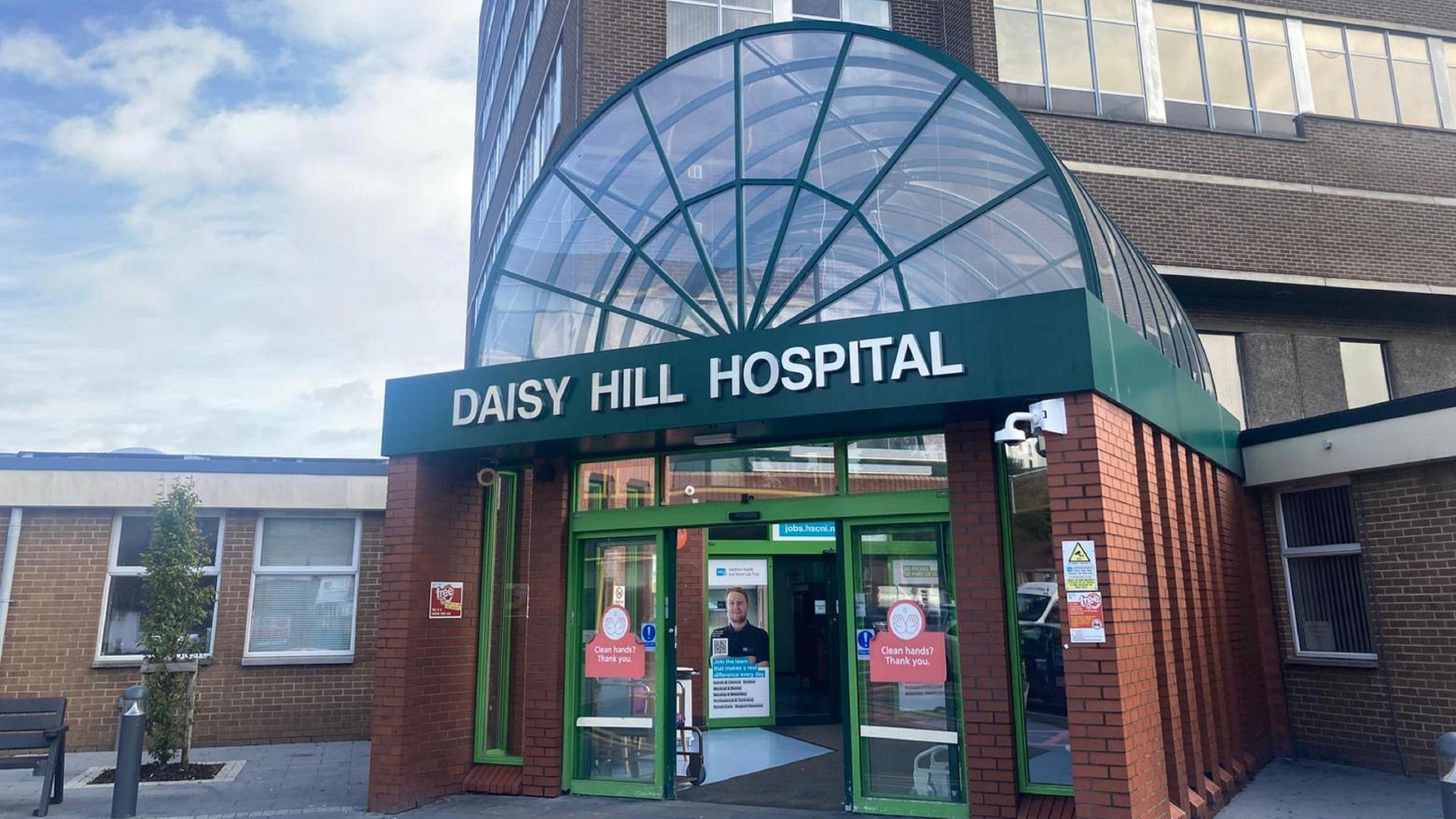 A large brown bricked building has a green banner on it's front that reads 'DAISY HILL HOSPITAL.' The banner sits at the entrance to the building below a glass archway which is encased in a green skeletal structure. They sit on-top of a red brick extension out the front of the brown bricked building. There is a black bench and a small green tree to the left of the entrance