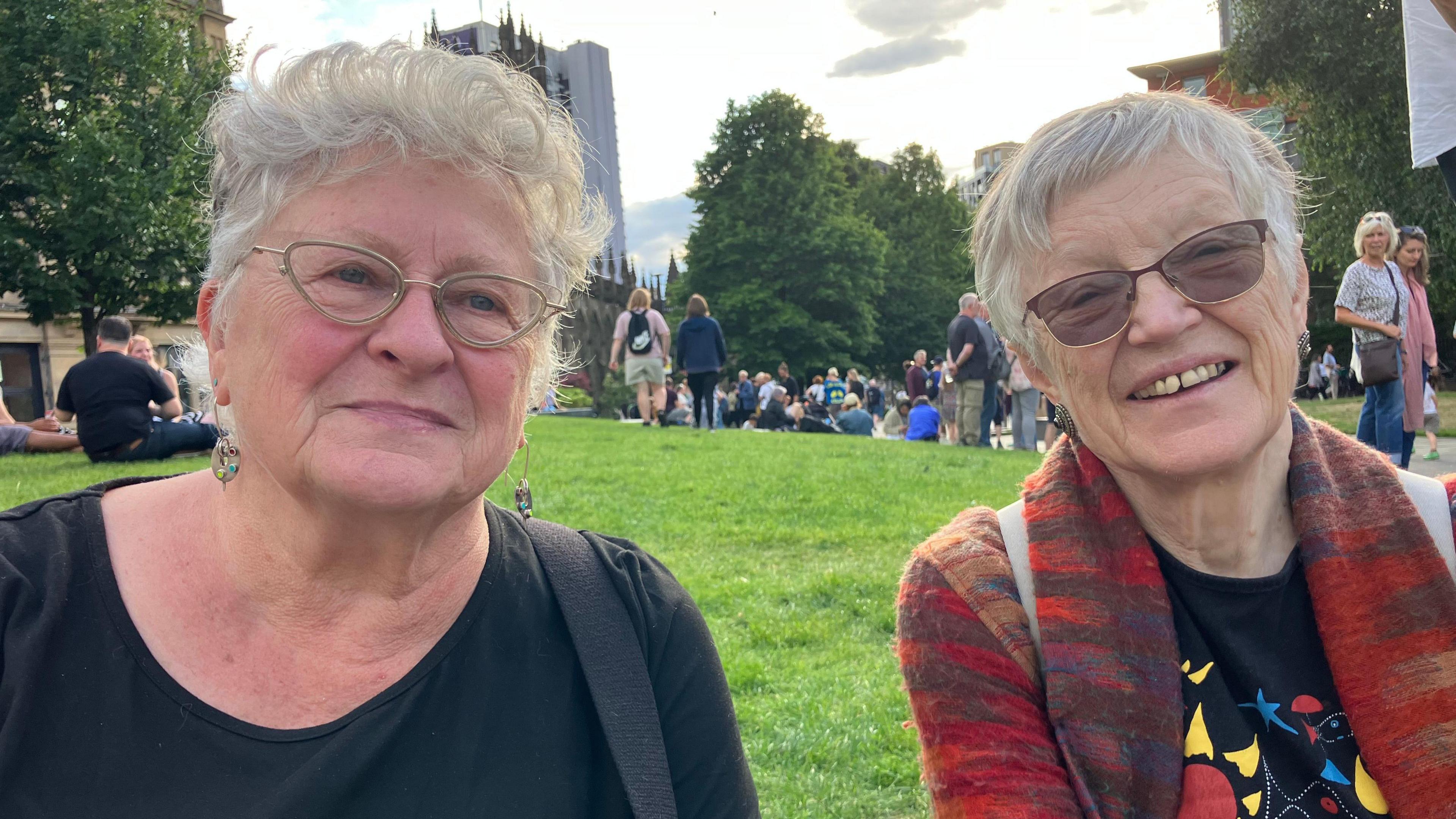 Two women with grey hair sit on the grass. They both have glasses. The woman on the left is wearing black. The one on the right is wearing red orange and blue cardigan and a black T-shirt with a picture of sunflower and fish on it.