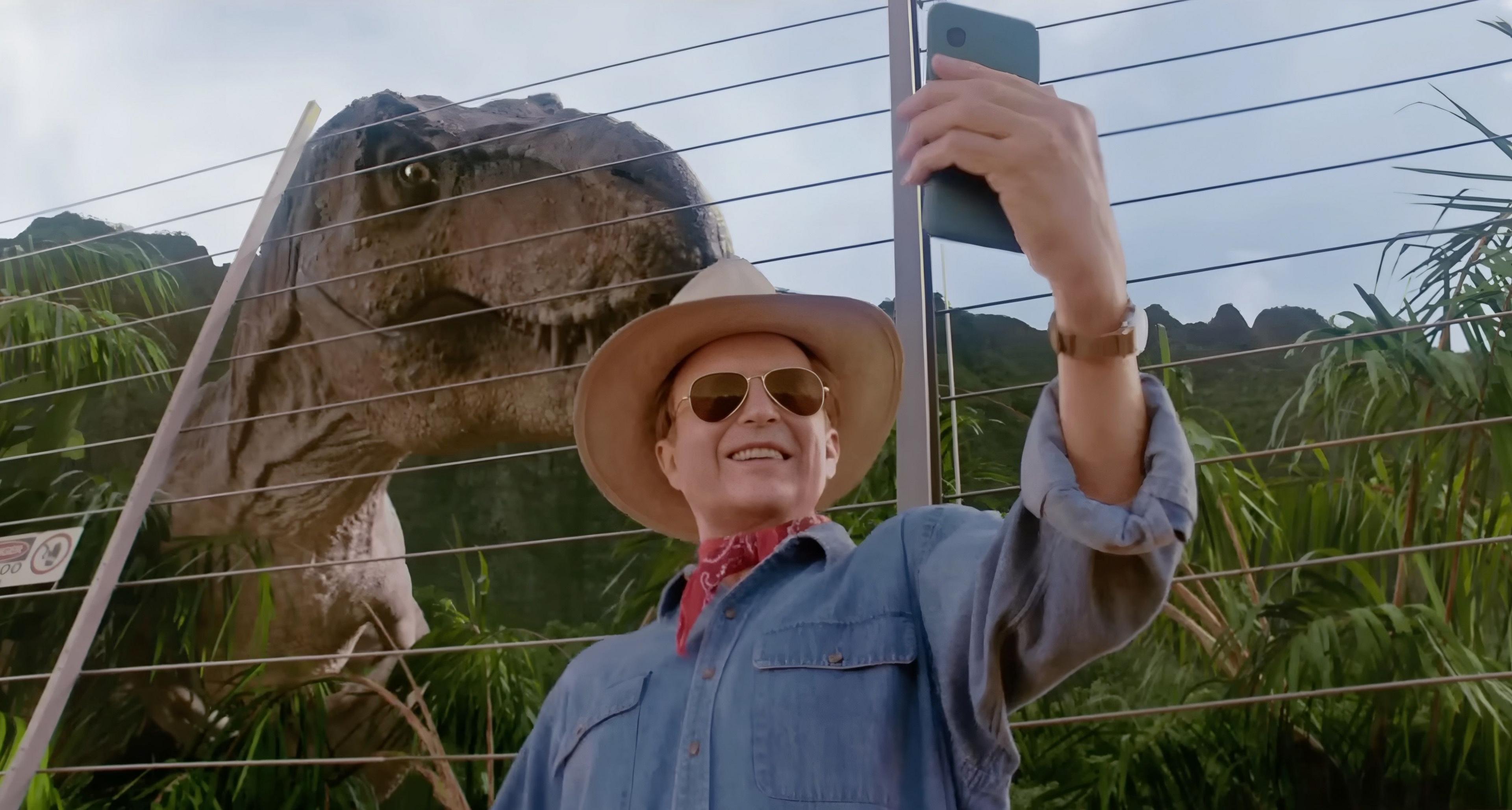 A man in a hat and sunglasses takes a selfie with a T.rex behind him in an enclosure. 