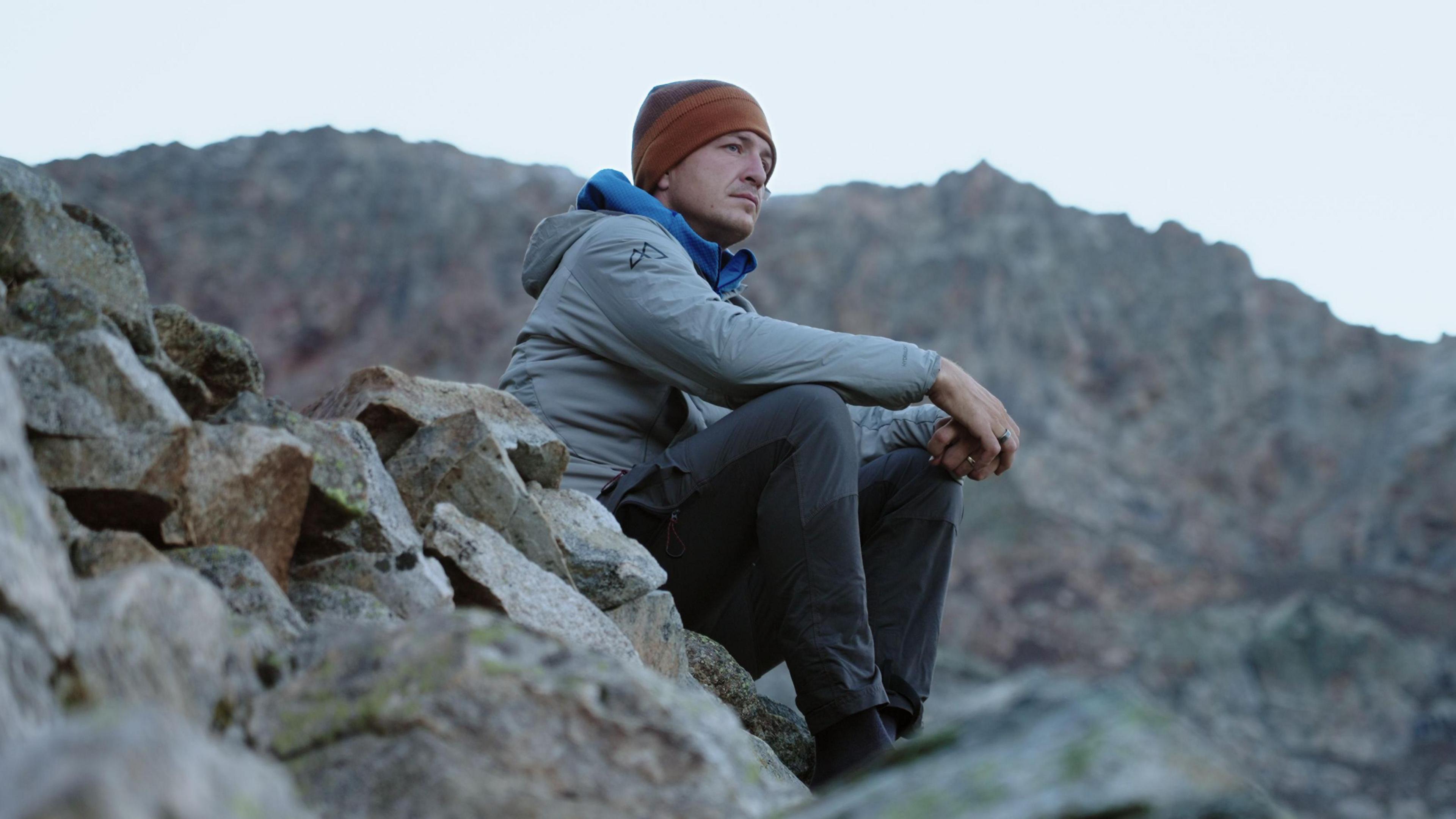 Ed Jackson sat on a rock. He is wearing an orange beanie and a grey jacket with dark trousers. He is looking out into the distance, with a blank expression on his face.