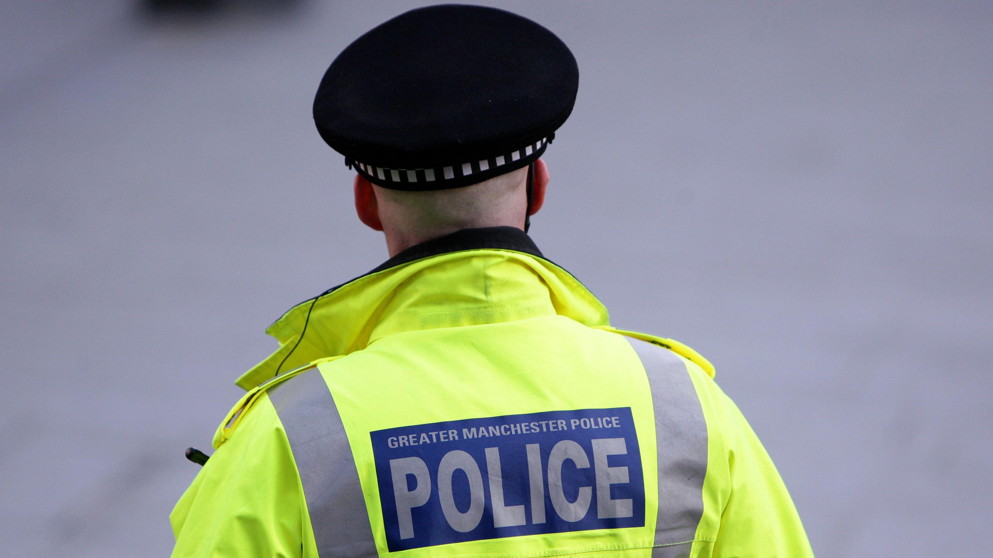 A police officer wears a hat and a hi-vis jacket with his back to the camera, a blue sign with white lettering, reads Greater Manchester Police, on his jacket