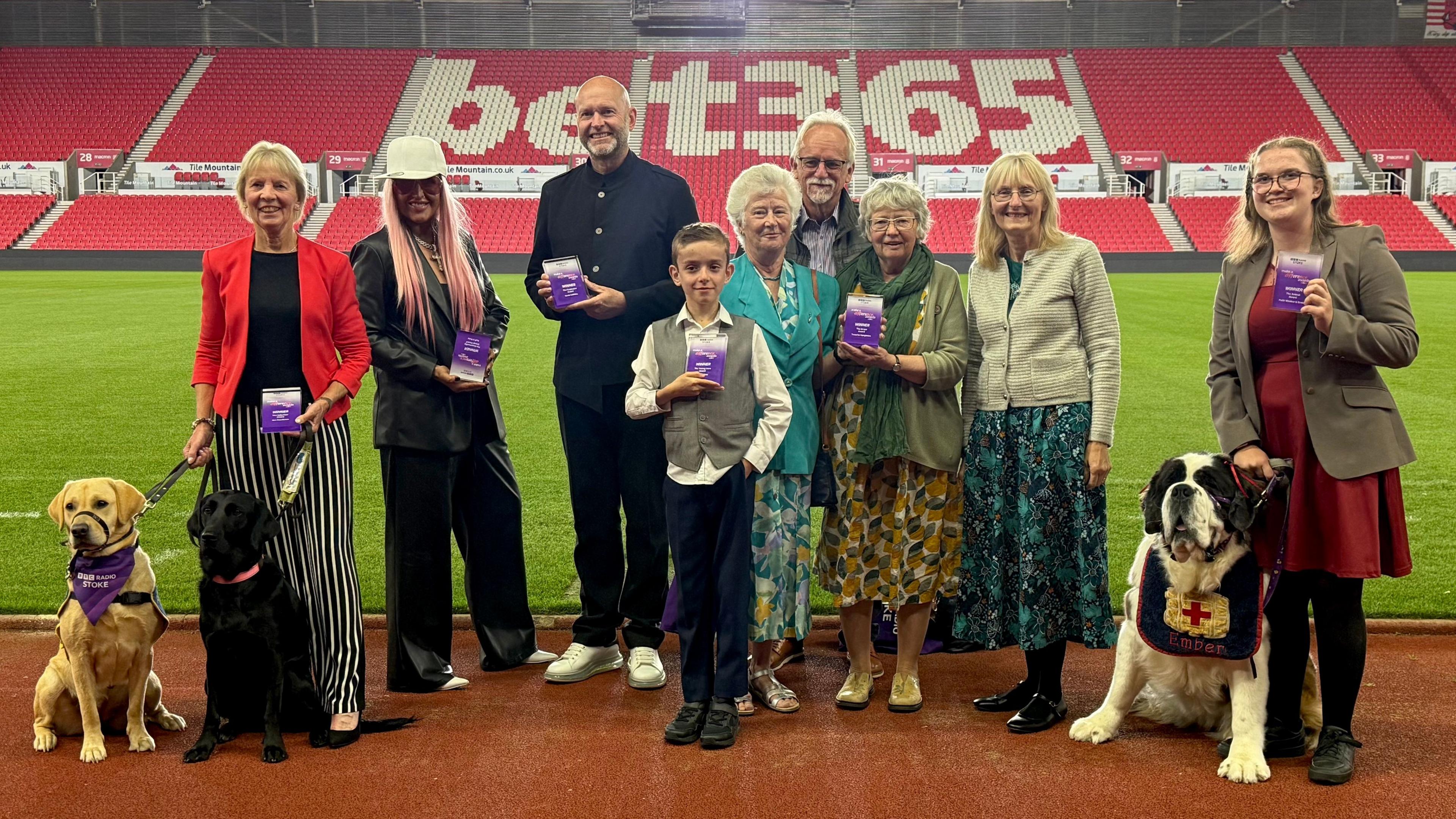 A group of winners from the BBC Radio Stoke Make A Difference Awards are gathered at the side of the pitch at the Bet365 Stadium. There is red branded Bet365 seating behind them and they are stood on the side of the football pitch.