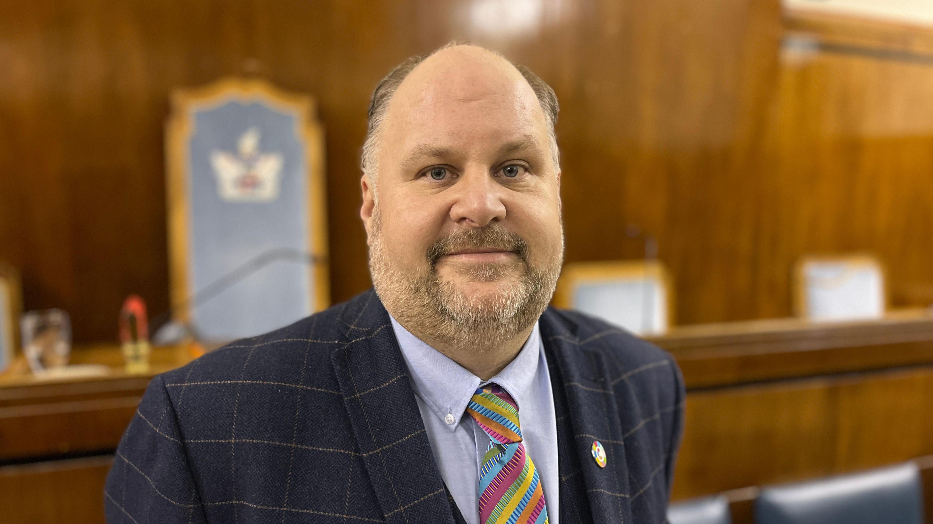 Councillor Jim Robbins looks into the camera, smiling slightly. He is wearing a blue suit, shirt and tie - the tie is colourful with stripes.