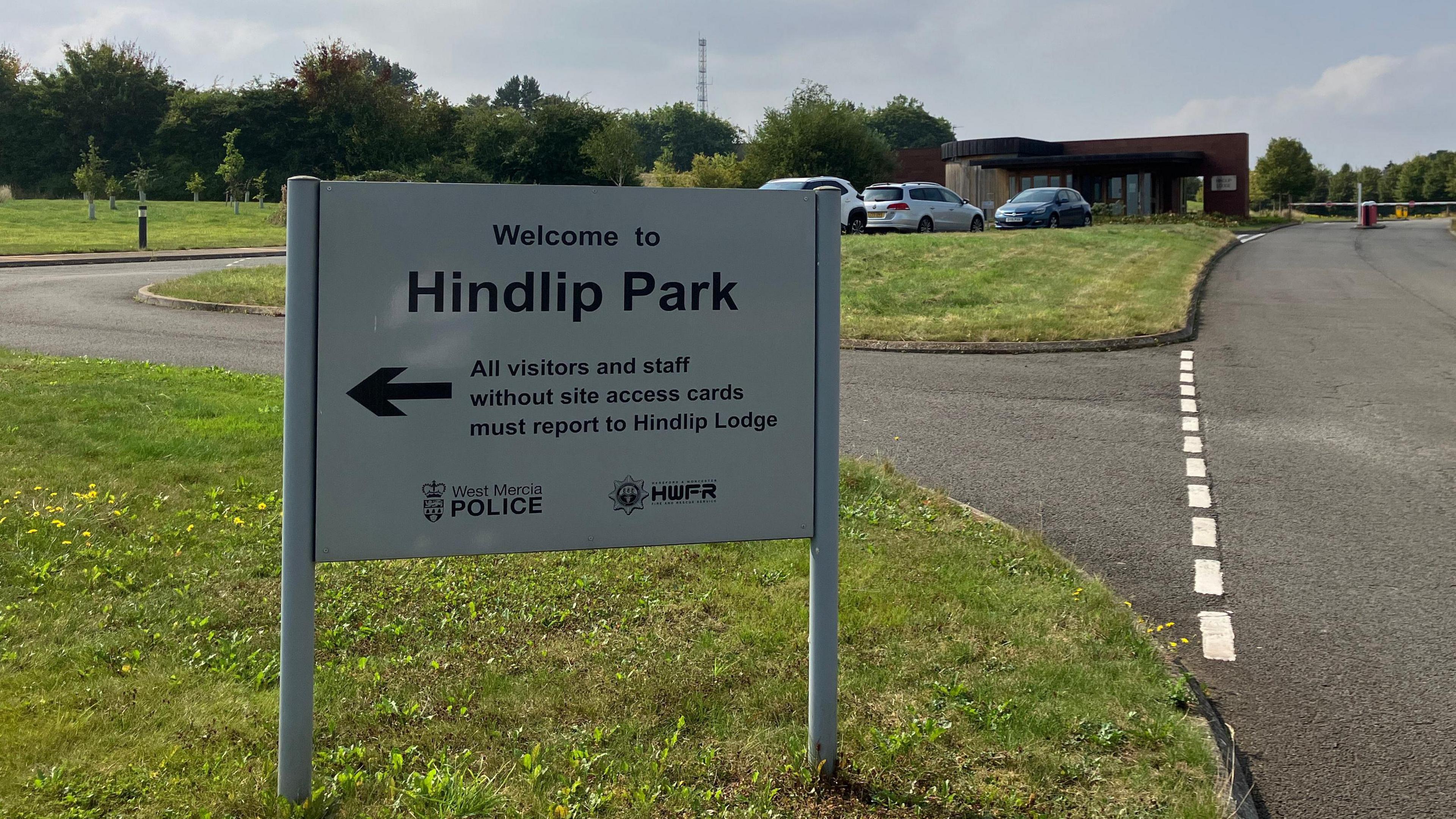 A sign at Hindlip Park, which is the headquarters of West Mercia Police, points visits and staff to the entrance. It is it a road junction surrounded by areas of grass. There are road markings and buildings and cars in the background.