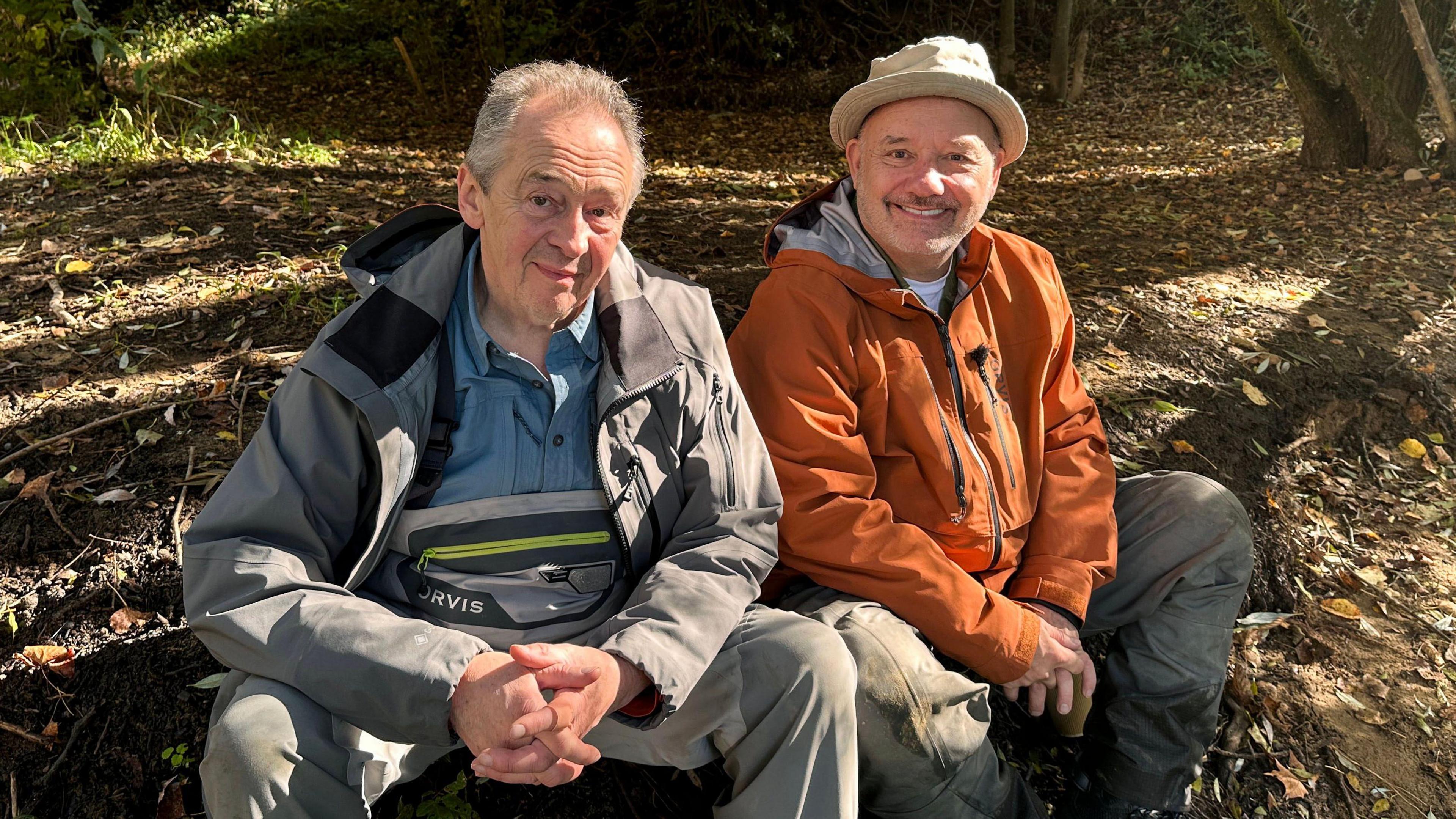 Paul Whitehouse with short, grey hair, wearing a grey coat and trousers has his hands clasped together as he sits down. Next to him is Bob Mortimer, wearing a beige hat and orange coat, and brown trousers, with his hands together sat down. They are surrounded by leaves on the floor in a forest. 