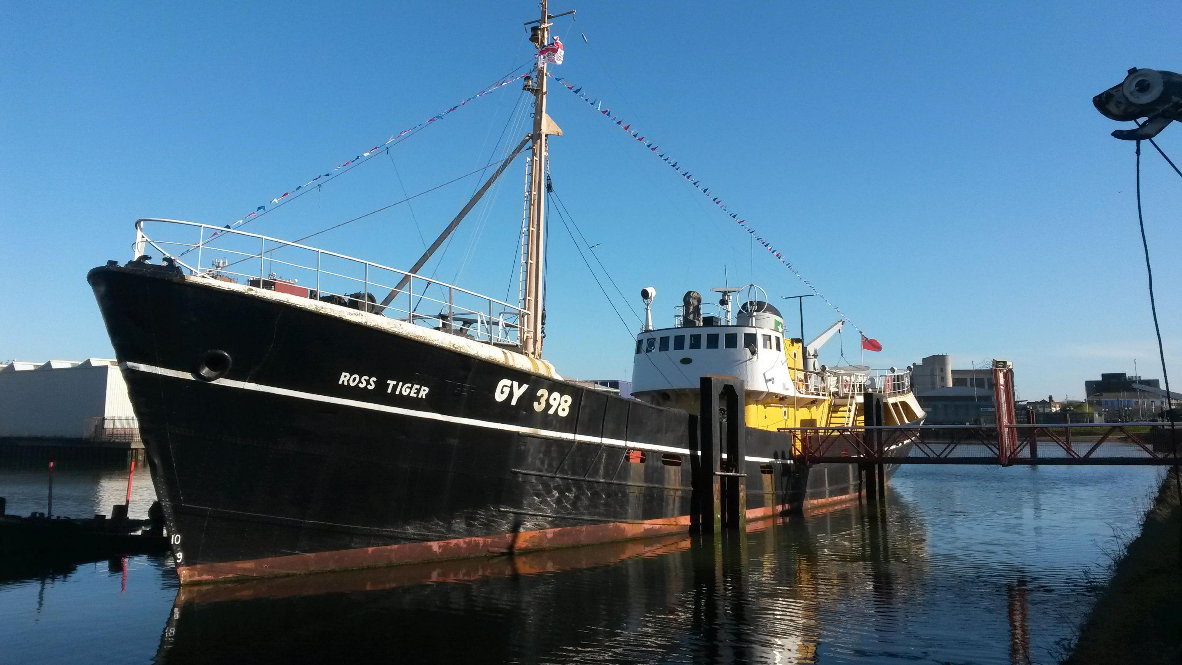 The Ross Tiger fishing trawler, sitting in its berth in the dock on a sunny day.  The boat has a large black hull and a white and yellow cabin and has multicoloured bunting up and down its rigging.