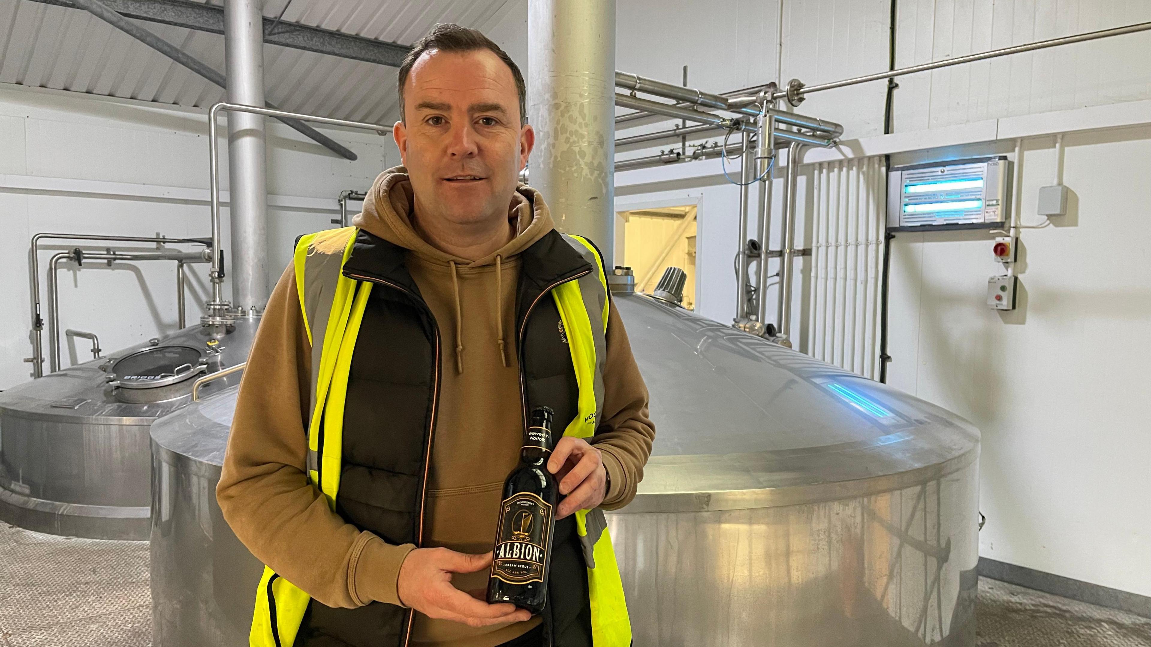 James Ramm, a man wearing a hi-vis jacket and a hoodie at a brewery. He is holding a bottle of stout in his hands.