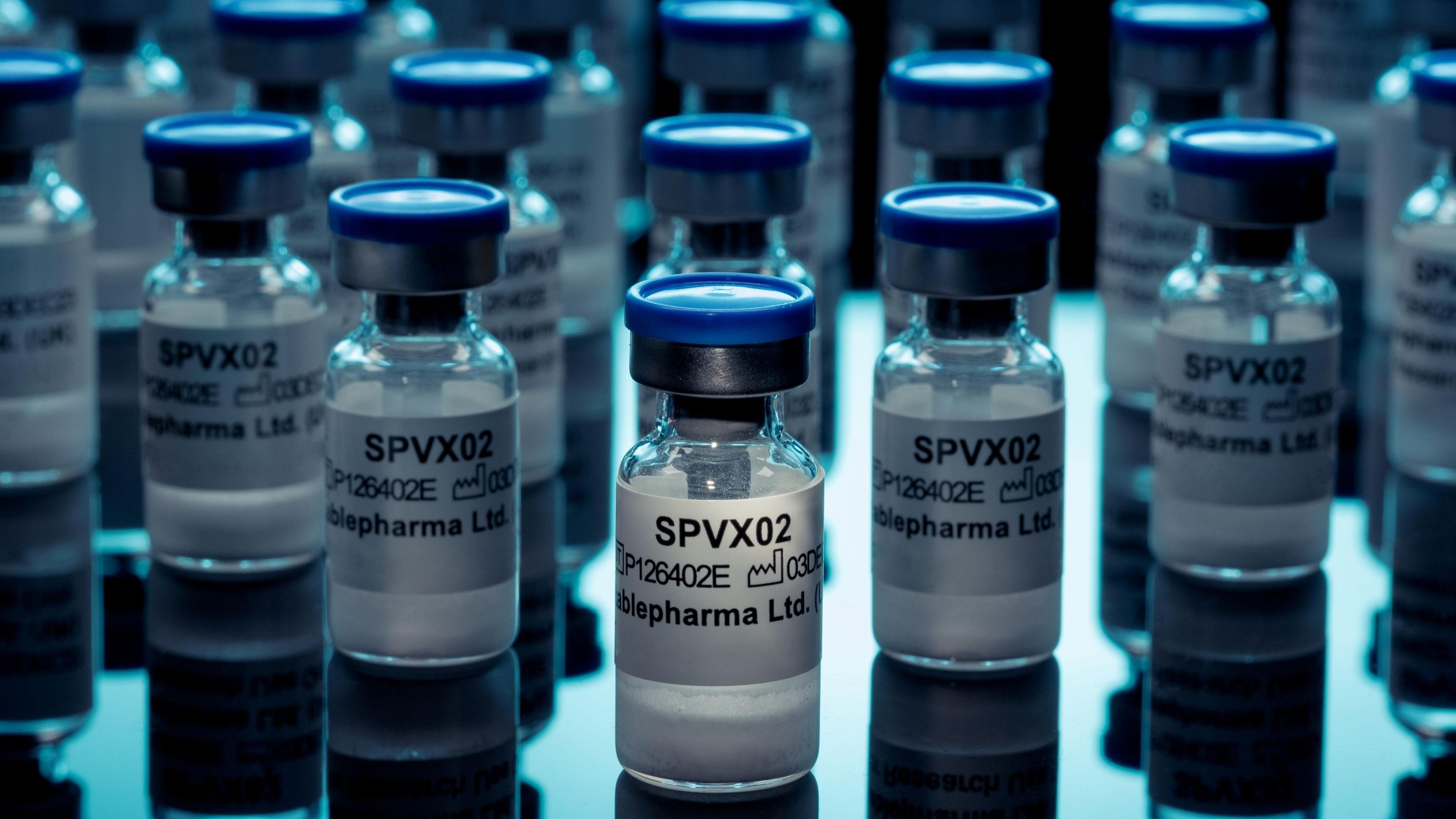 This image shows multiple small glass pharmaceutical vials arranged on a reflective surface under cool blue lighting. The vials have blue caps and white labels, with the text “SPVX02” clearly visible on several containers; additional smaller text appears to include a product code and the company name Stablepharma Ltd. The composition places one vial in sharp focus near the center foreground, while numerous similar vials extend into the background with progressively softer focus. The vials appear to contain a white powder. Reflections of the vials are visible on the glossy surface beneath them, creating a symmetrical, laboratory-style aesthetic. The color palette is dominated by blues, whites, and blacks, and the lighting emphasizes the glass, metal, and plastic textures of the containers. The overall setting resembles a pharmaceutical, biotechnology, or medical research environment, with the image focusing on the repeated pattern of labeled drug or vaccine vials.