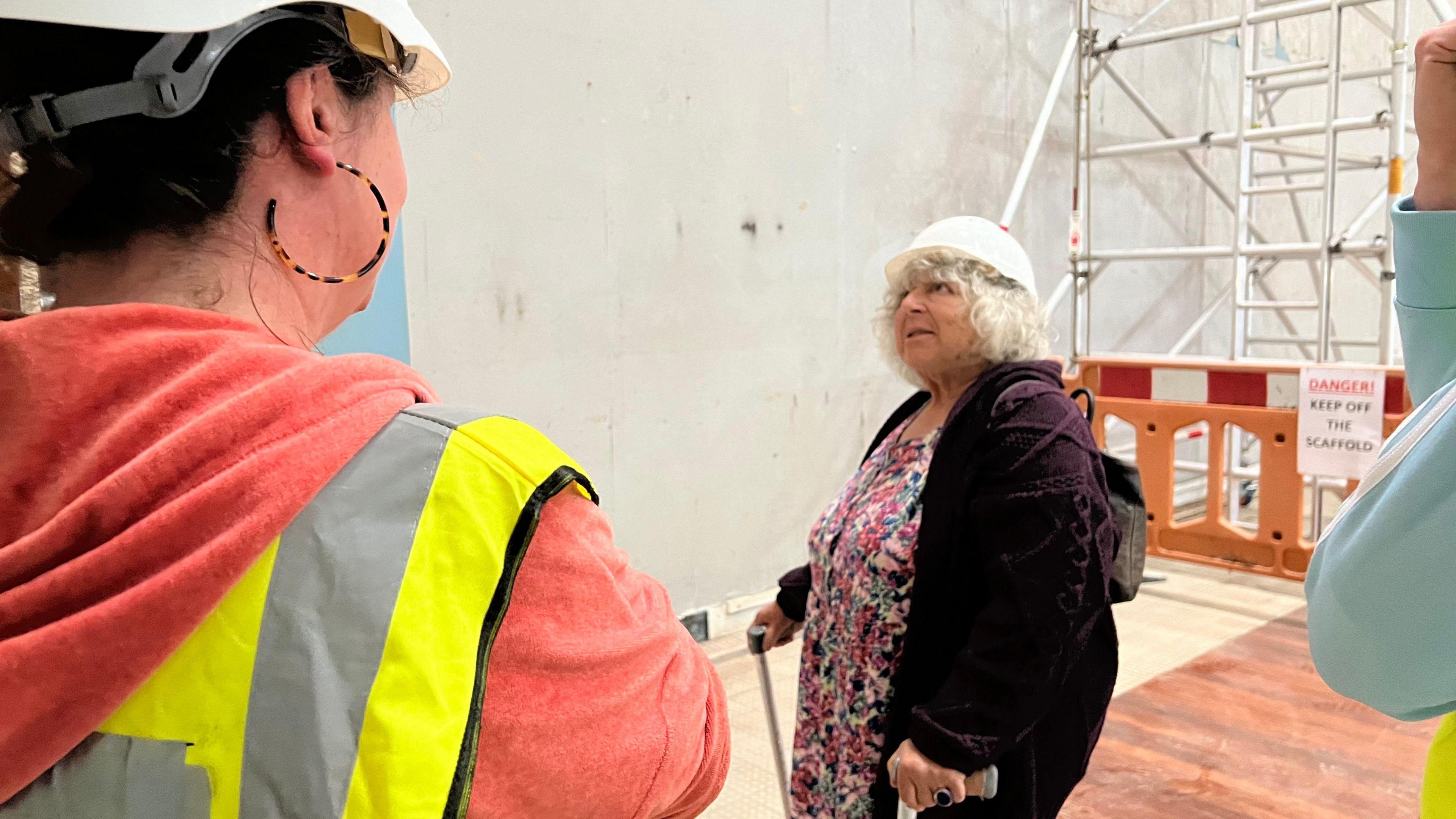 Miriam during a visit to the site. She wears a white hard hat, a pink floral dress and a purple jumper. Other people in high-vis jackets can be seen in the foreground watching Miriam. 