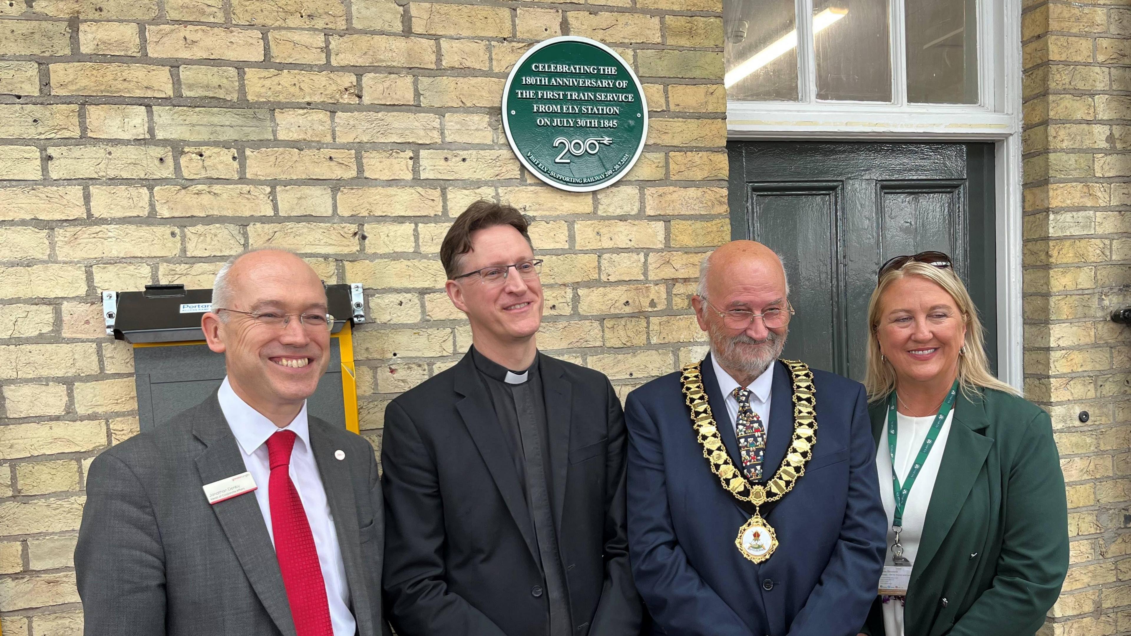 Four people on the platform of a railway station including the Mayor of Ely, wearing a ceremonial gold chain and the Canon of Ely Cathedral wearing a dog collar.