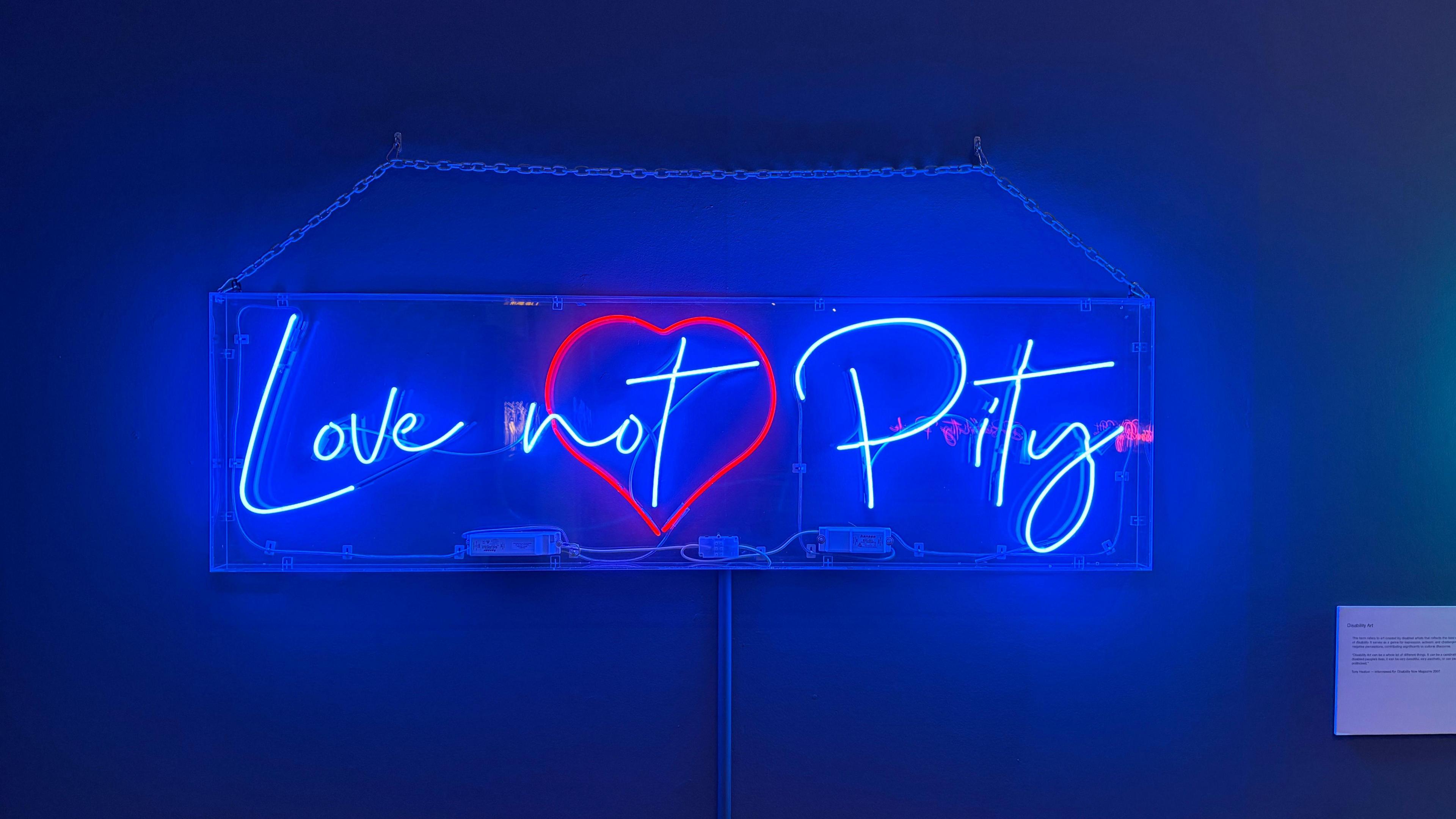 A blue neon sign with a red heart in the middle of it. The writing reads "love not pity"