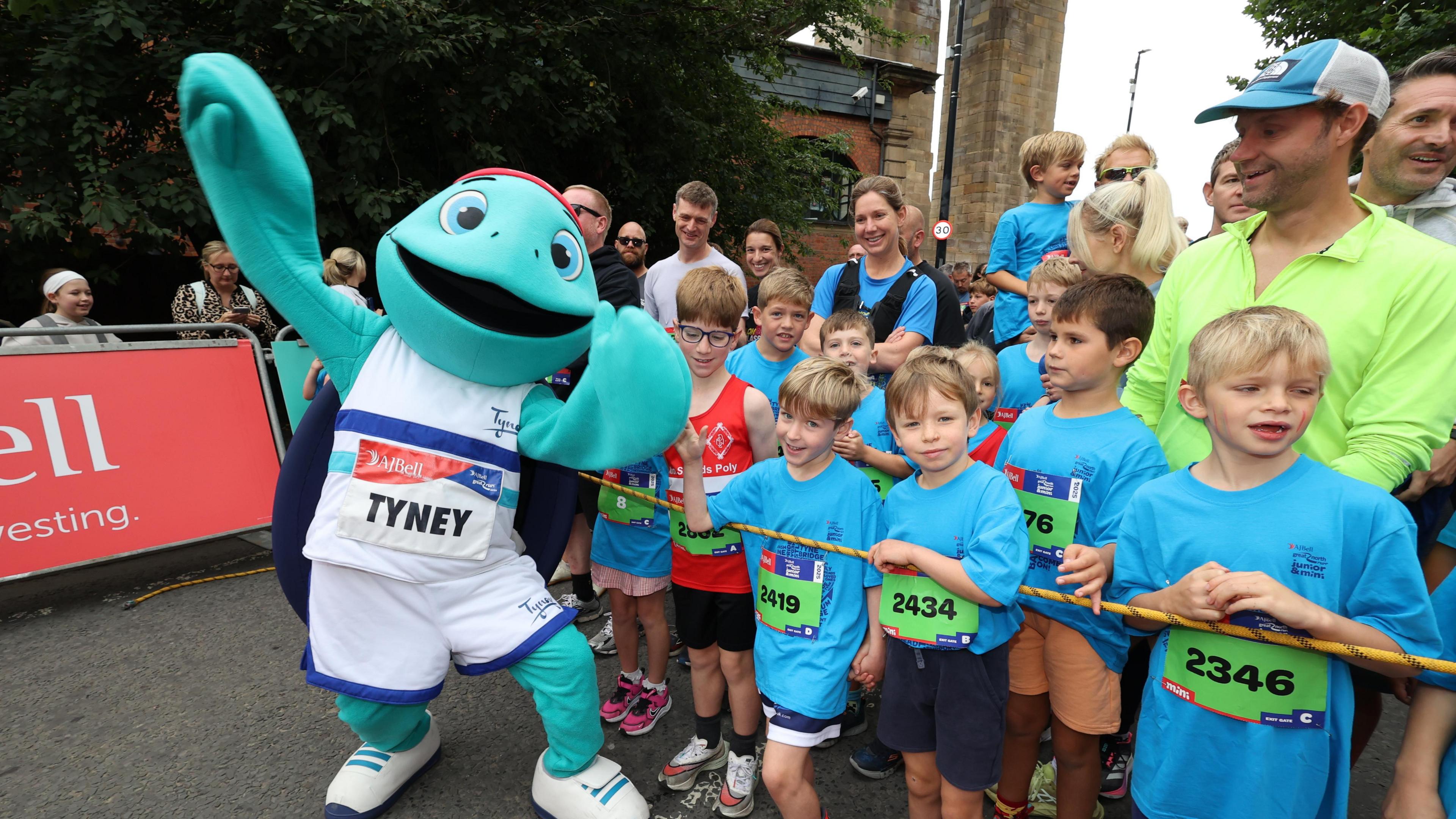 Man in life-sife green turtle costume poses for the camera with their arms in the air. He is next to a group of children who are all wearing blue T-shirts and green signs on their fronts with their race numbers. They are standing behind a rope. Parents stand in the background looking happy.