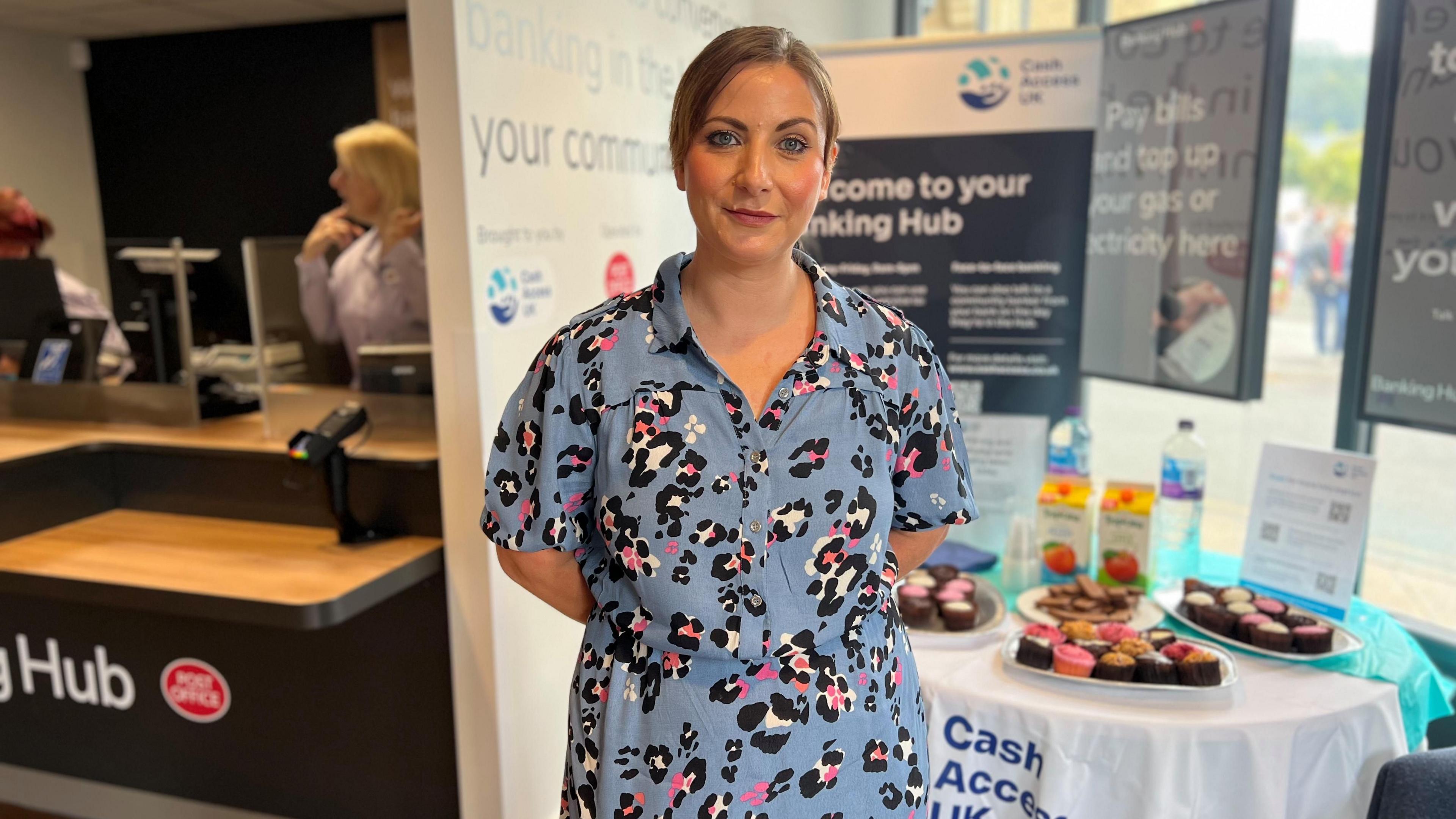 Woman with brown hair tied up in light blue dress with pink and white animal print stood infront of bank service counter and table with cakes on