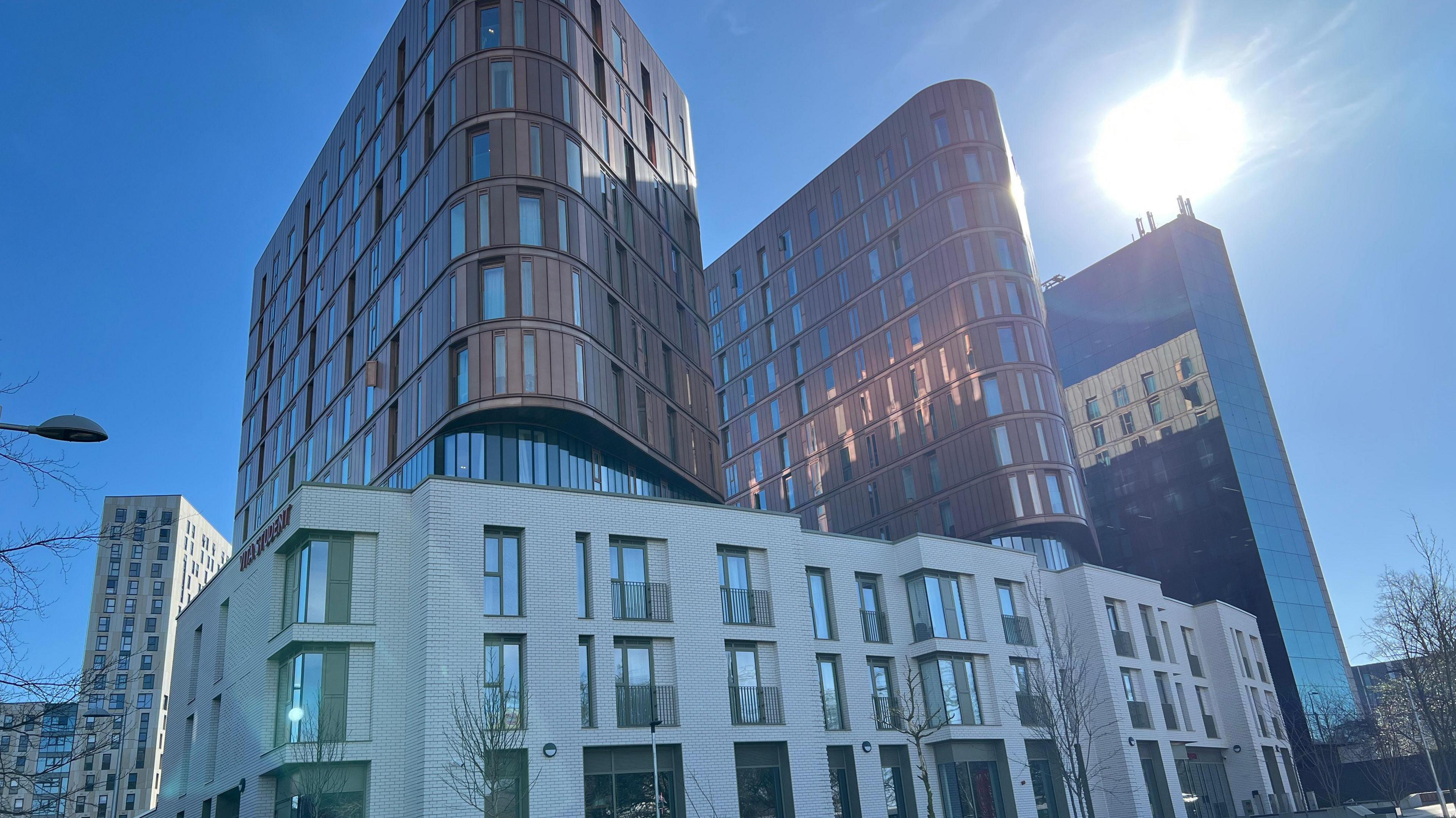 University accommodation building, grey with rectangular windows and a brown building with two curves above this. The backdrop is of blue sky and sun.