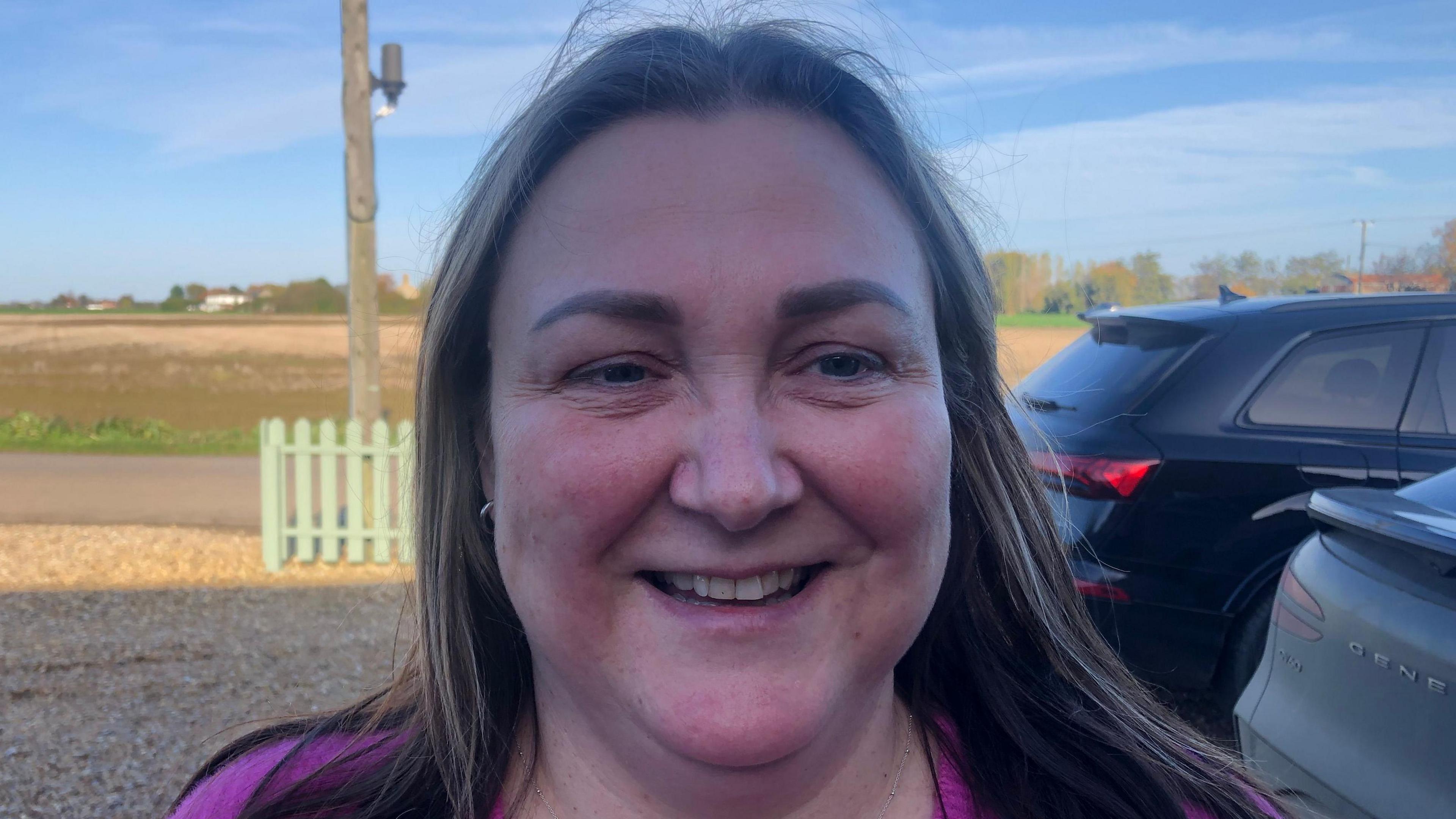 Karen Baldwin has sandy coloured, shoulder length hair parted in the middle at the top of her forehead. She is wearing a pink 'V' necked jumper and is smiling. Behind her is farmland, with ploughed light brown soil visible. There are two cars parked on the right on a gravel drive way and front garden. A light green picket fence can be seen on the perimeter of her property.