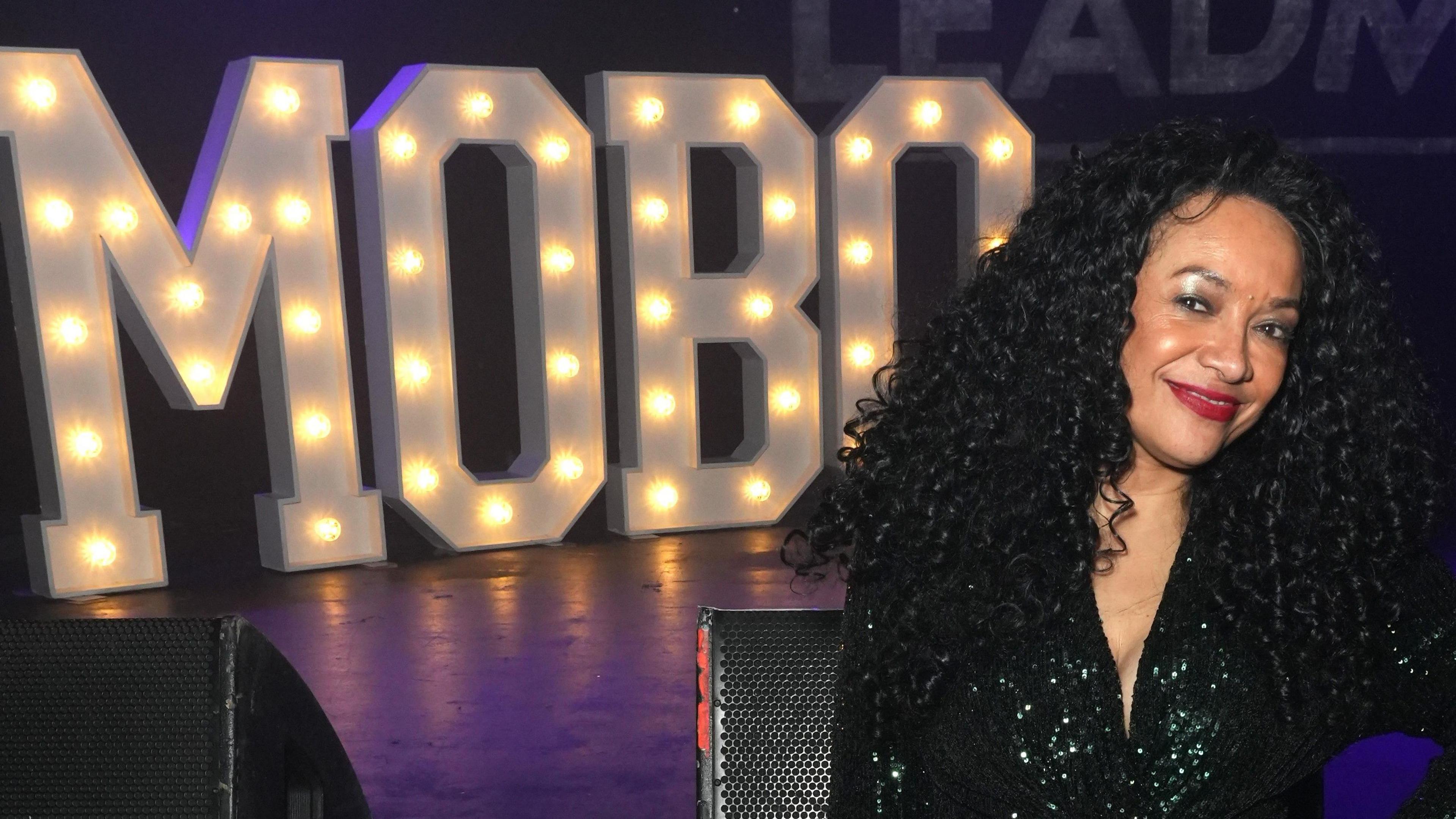 Kanya King at the 2024 MOBO Awards. Kanya has her hand on her hip and smiles at the camera, wearing a sparkly black dress. Behind her is a stage with sound equipment and MOBO spelled out in big lights. 