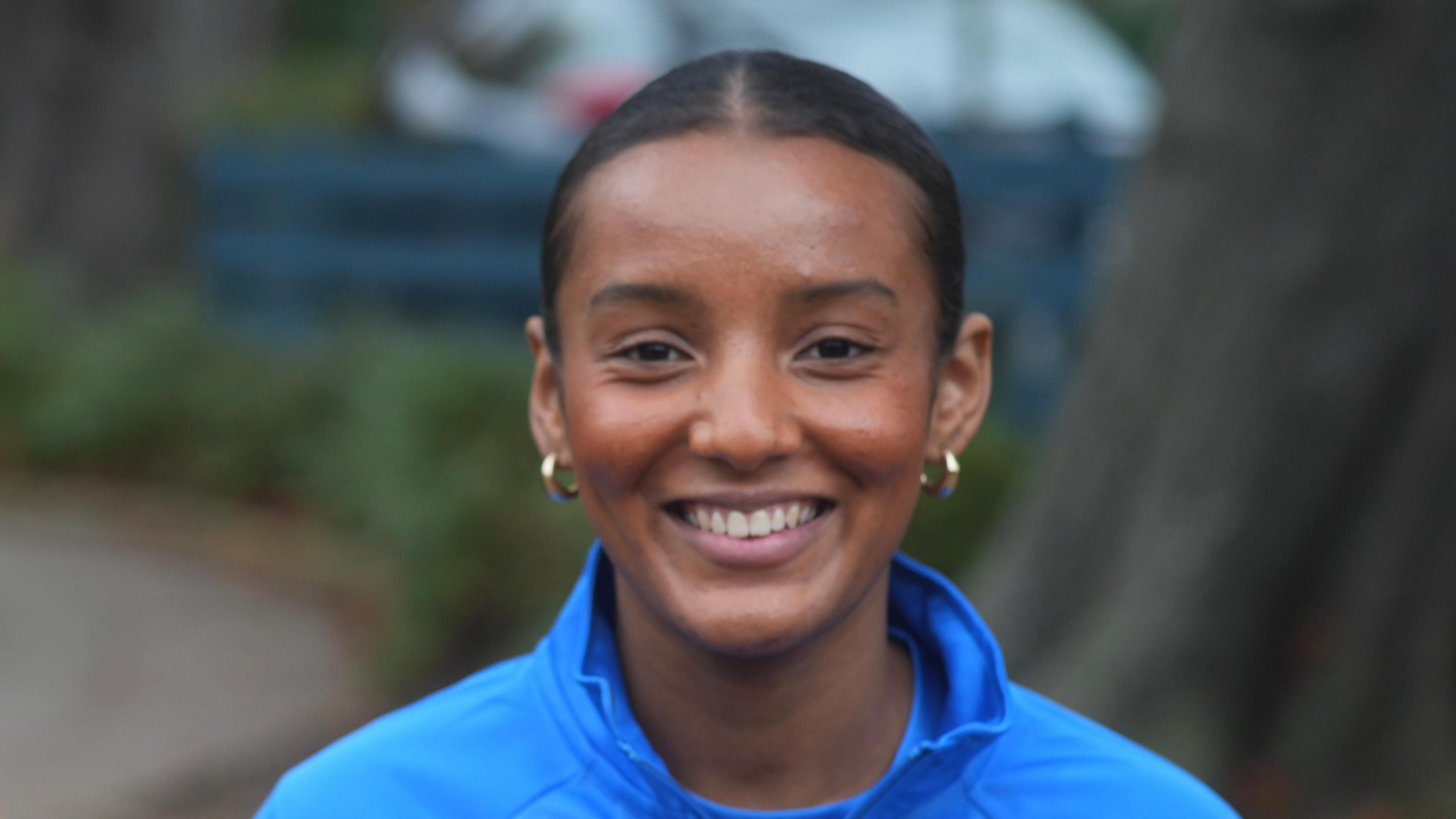 Justice Hall is smiling at the camera. Her dark hair is tied up behind her head. She is wearing gold hoop earrings and a blue jumper with a blue top on underneath.