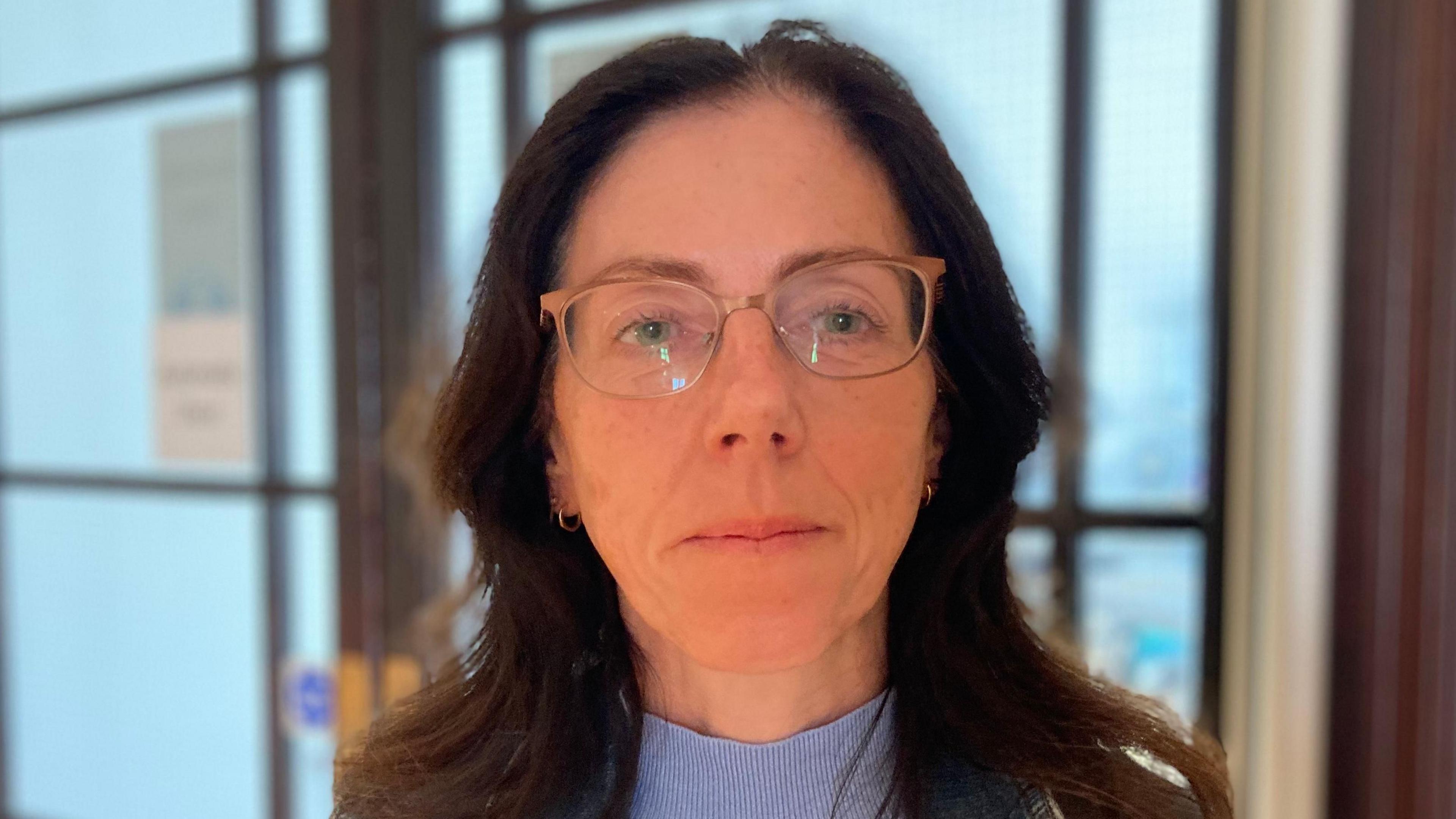 A woman with brown hair is looking into the camera. She wears glasses and is wearing a blue jumper and gold earrings. 