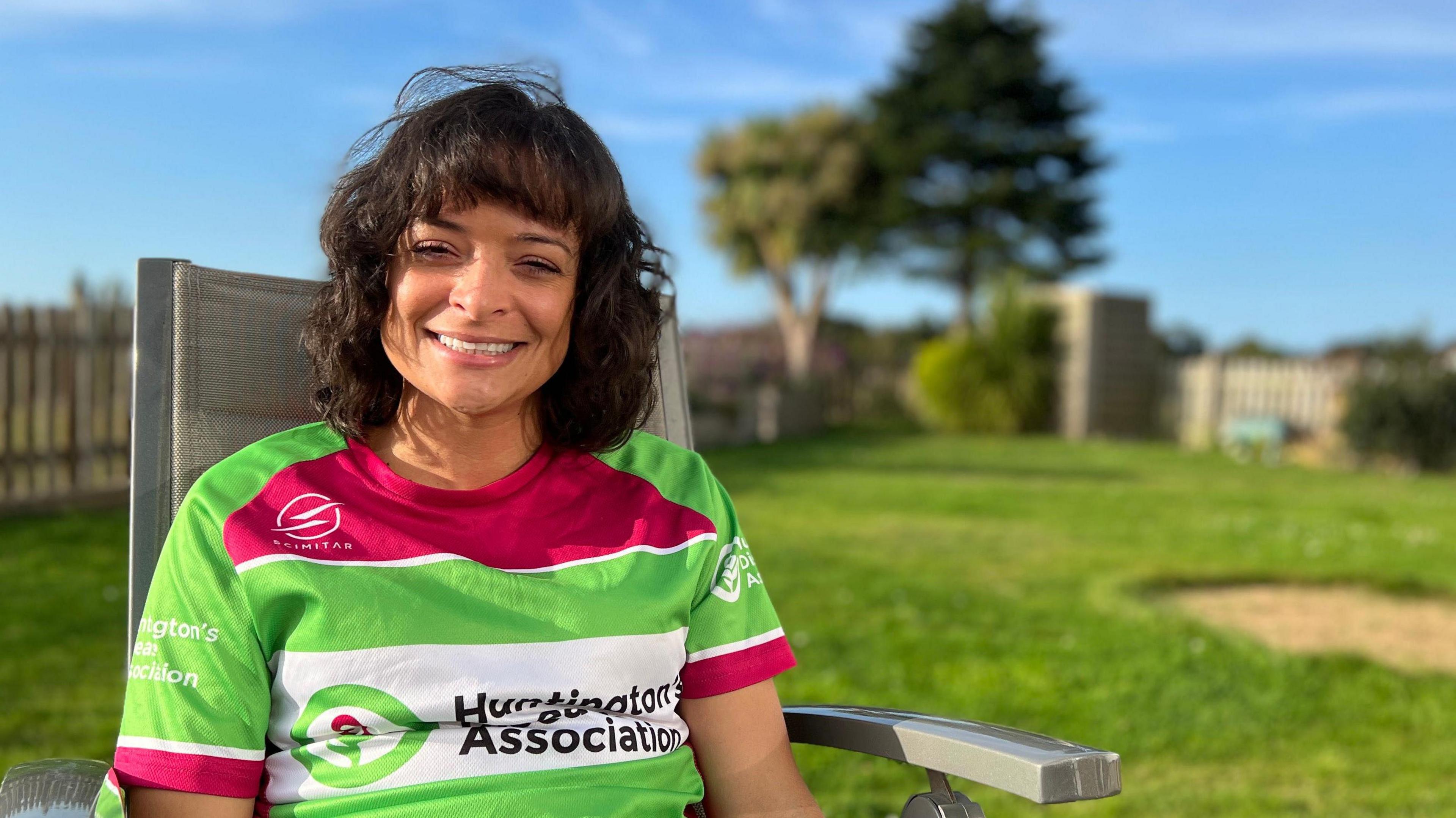Ashley has curly brown hair is smiling while sitting on a chair outdoors in a grassy backyard. She is wearing a colorful sports jersey that reads "Huntington's Association" with green, pink, and white stripes. The background features a wooden fence, trees, and a house under a clear blue sky.