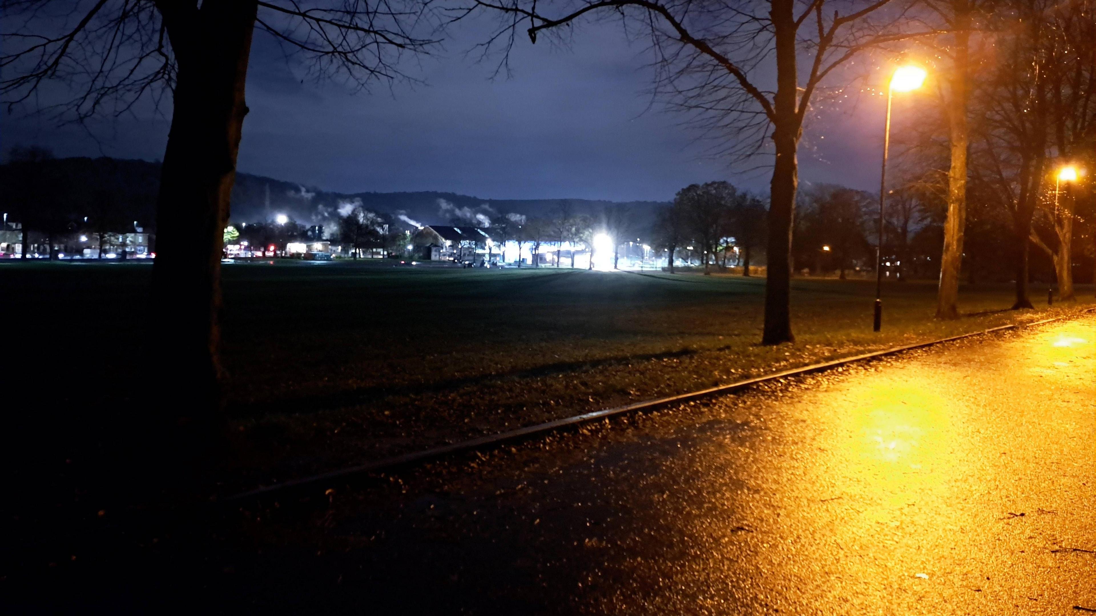 A view of a park at night shows a yellow glow from a street lamp while the rest of the park is in complete darkness with some house lights in the distance