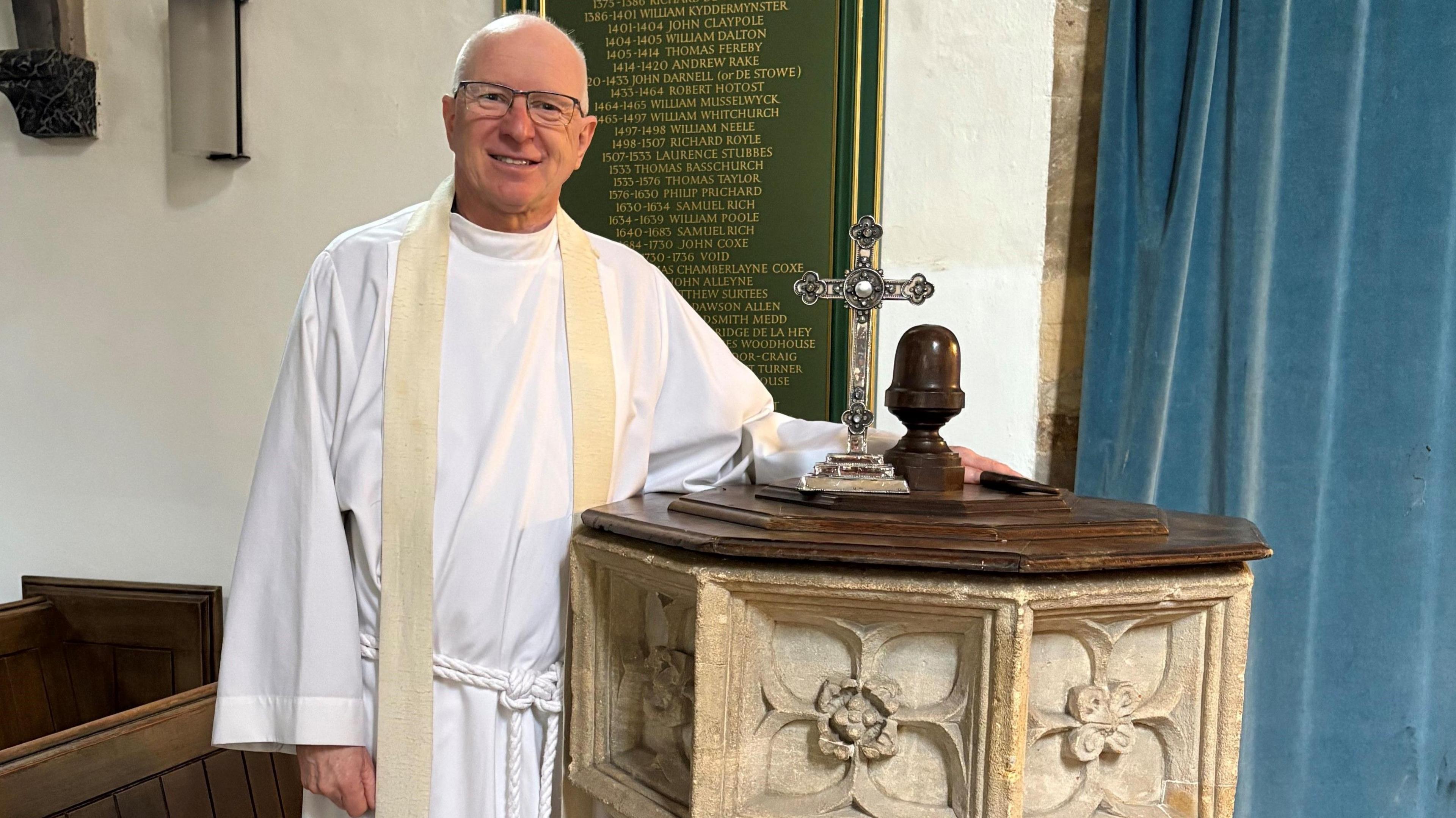 The Reverend David Minns posing alongside the cross, which is sitting on top of a hexagonal stone altar. He is wearing long white robes, has short white hair and glasses on. 