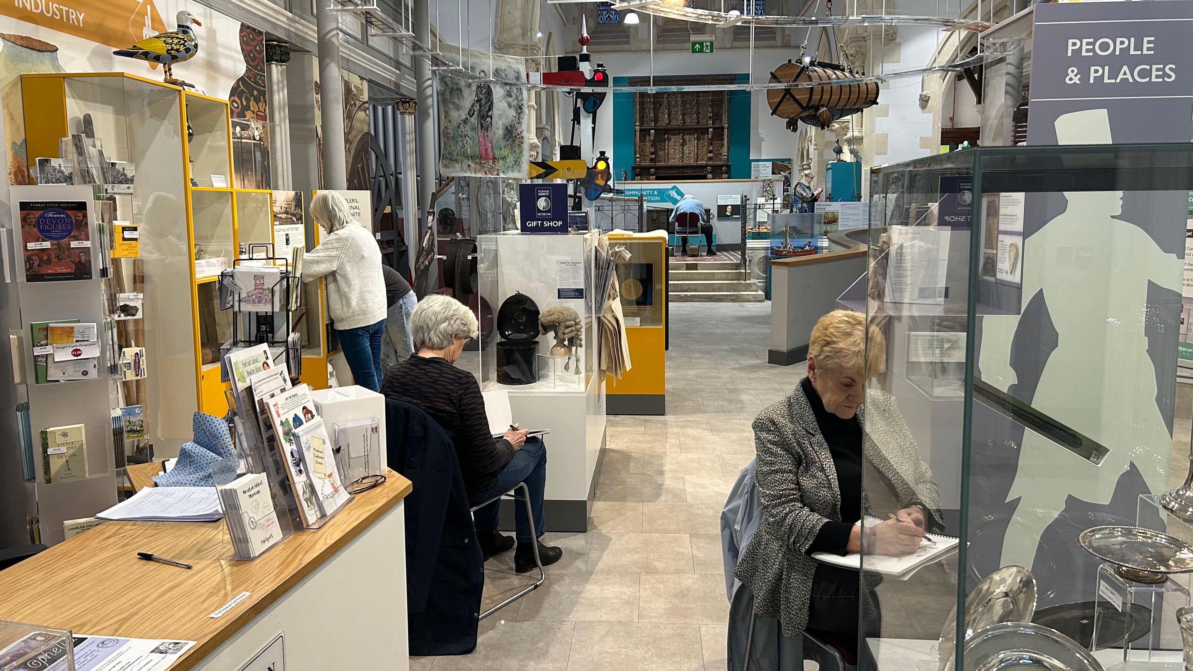 Three women sit with sketch books in front of display cases containing a range of artefacts