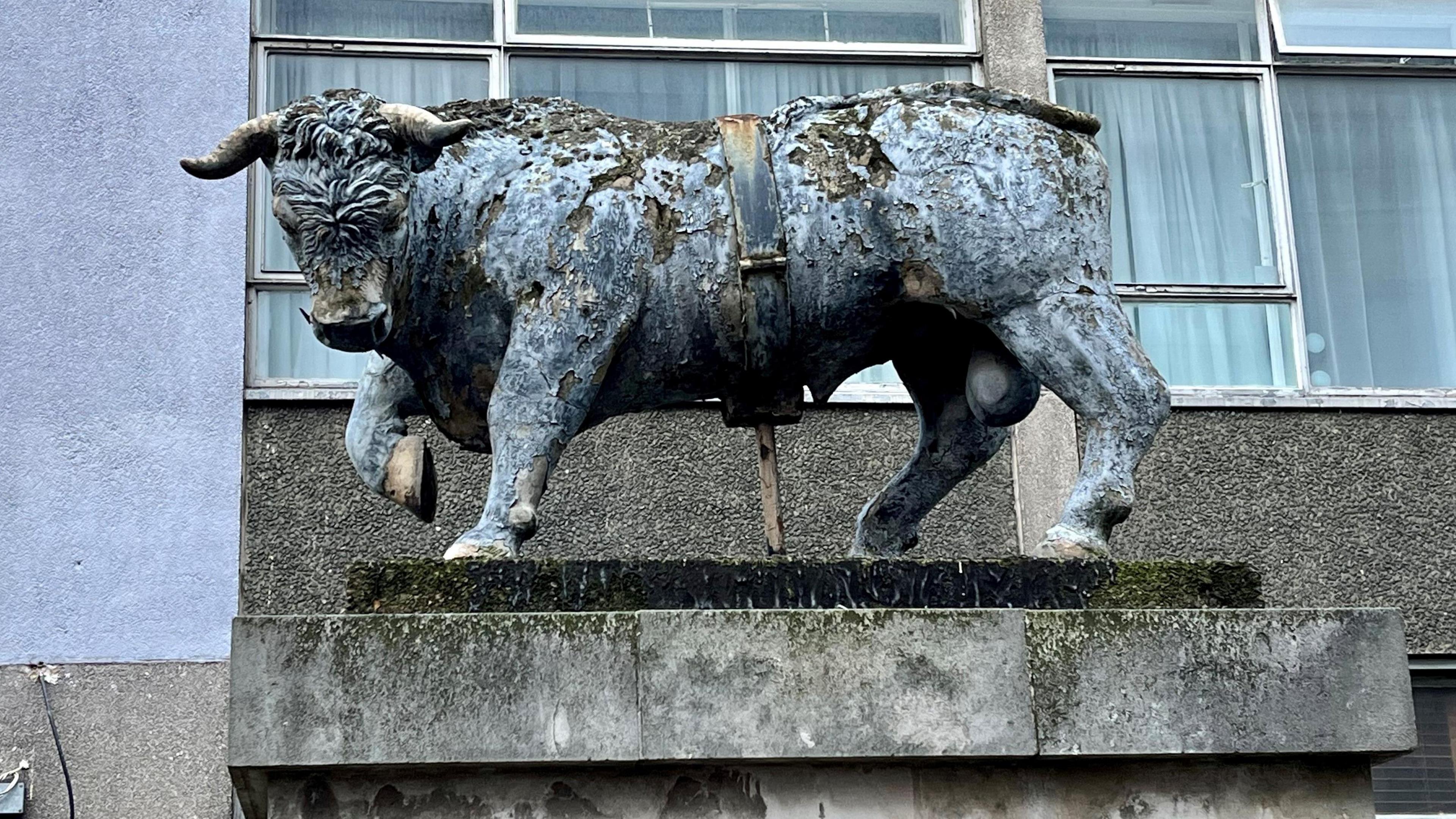 A large blue bull with grey horns and hoofs stands on a stone plinth with the windows of a hotel behind it