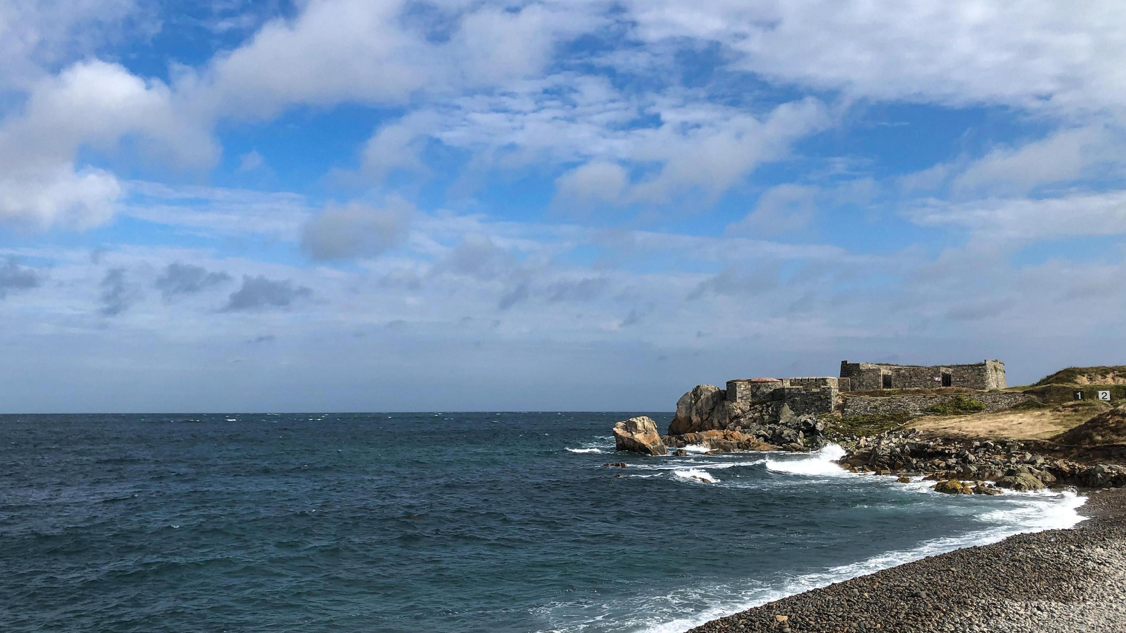 view of a shingle beach with what appears to be historical ruins on the headland. Sunny spells in the skies above