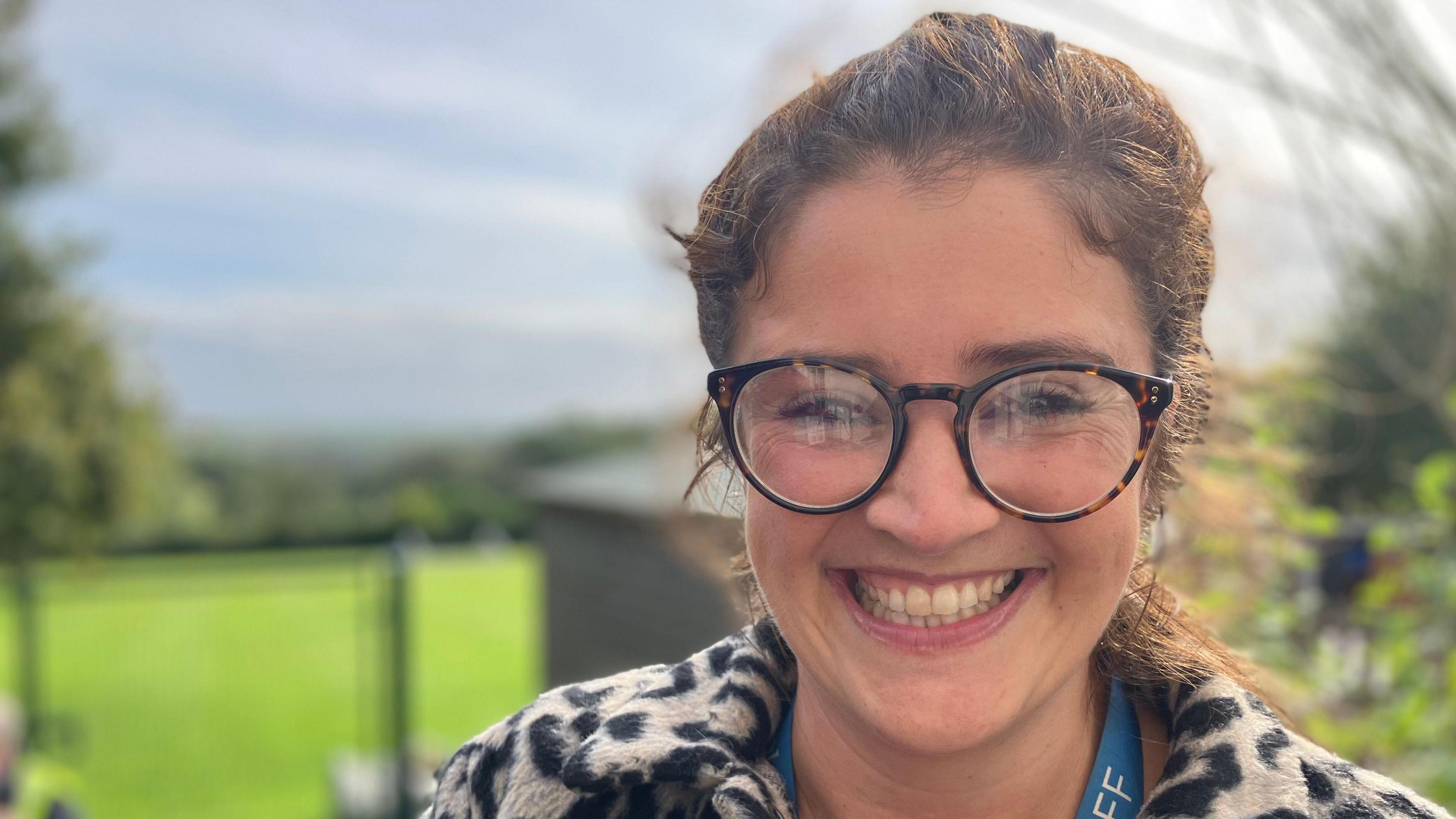 A smiling woman with glasses and a lanyard saying "Staff" stands in front of a field.