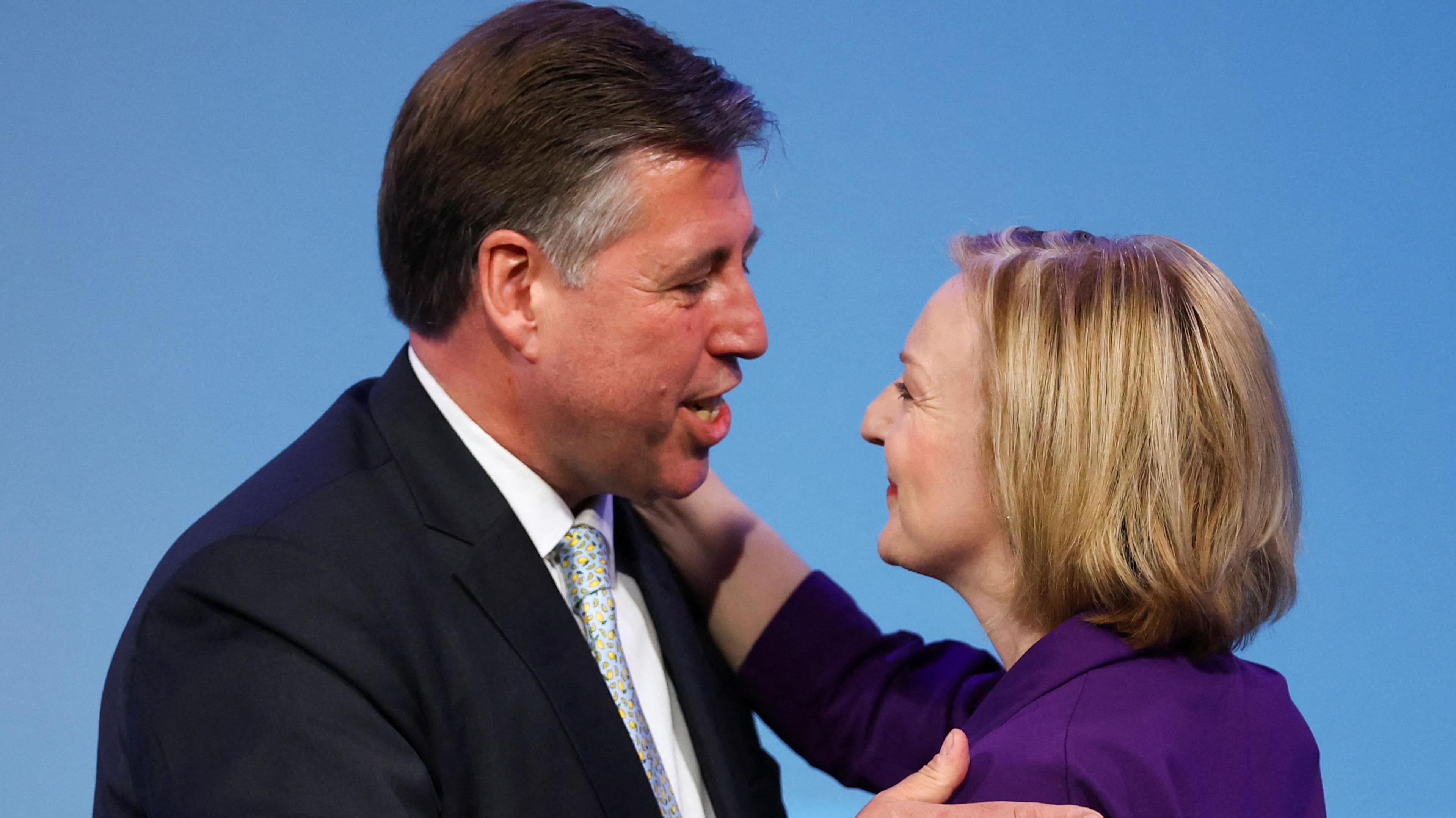 Lord Graham Brady, wearing a dark blue suit, white shirt and light blue tie, embraces a smiling Liz Truss, who has shoulder length blonde hair and is wearing a purple suit