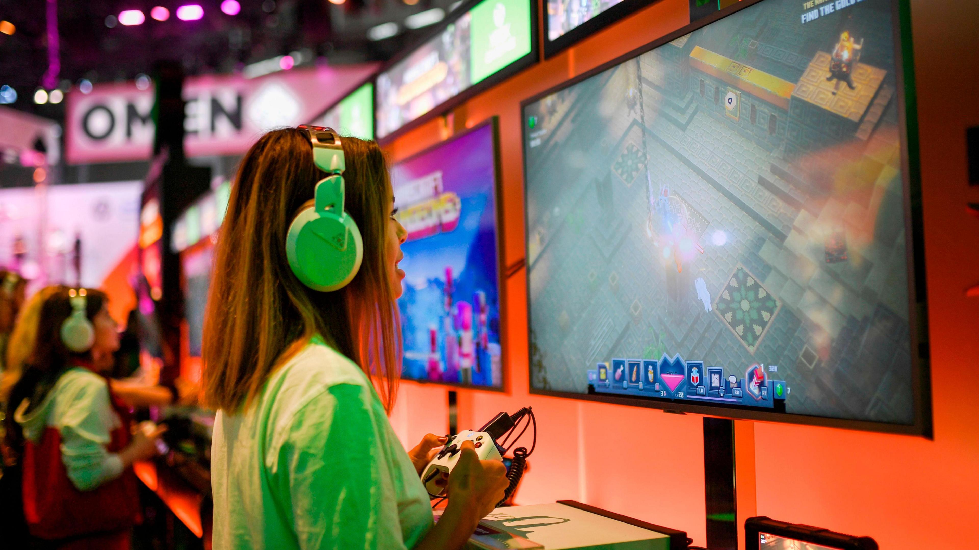 A woman wearing a white T-shirt and over the ear headphones plays on the Xbox at a stand at a video games trade fair. She's holding the controller and looking at a large screen in front of her.