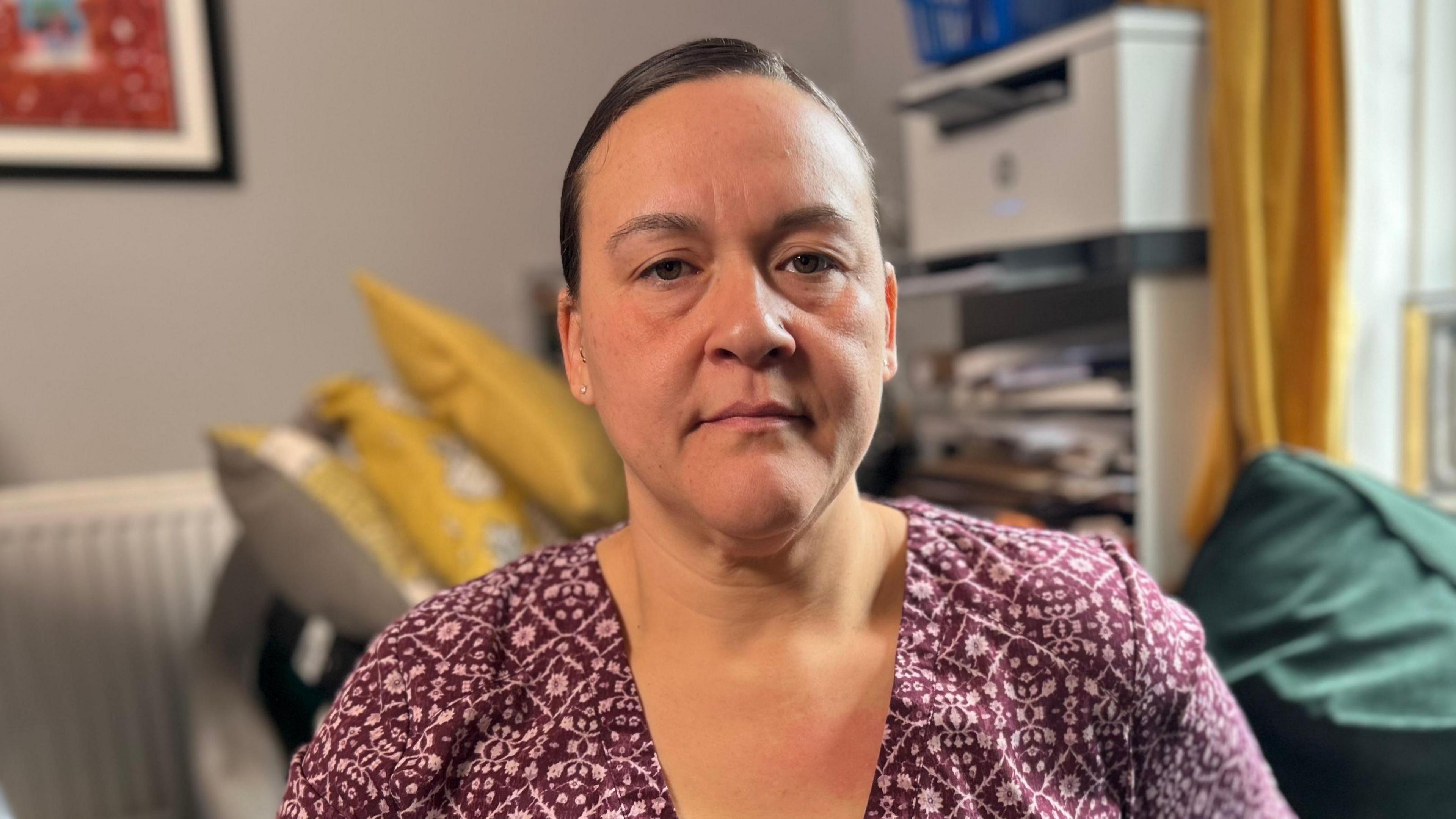 Jodi Jenny, wearing a purple coloured dress with a v-neck looks pensively at the camera as she sits in her home. There is a sofa behind her.