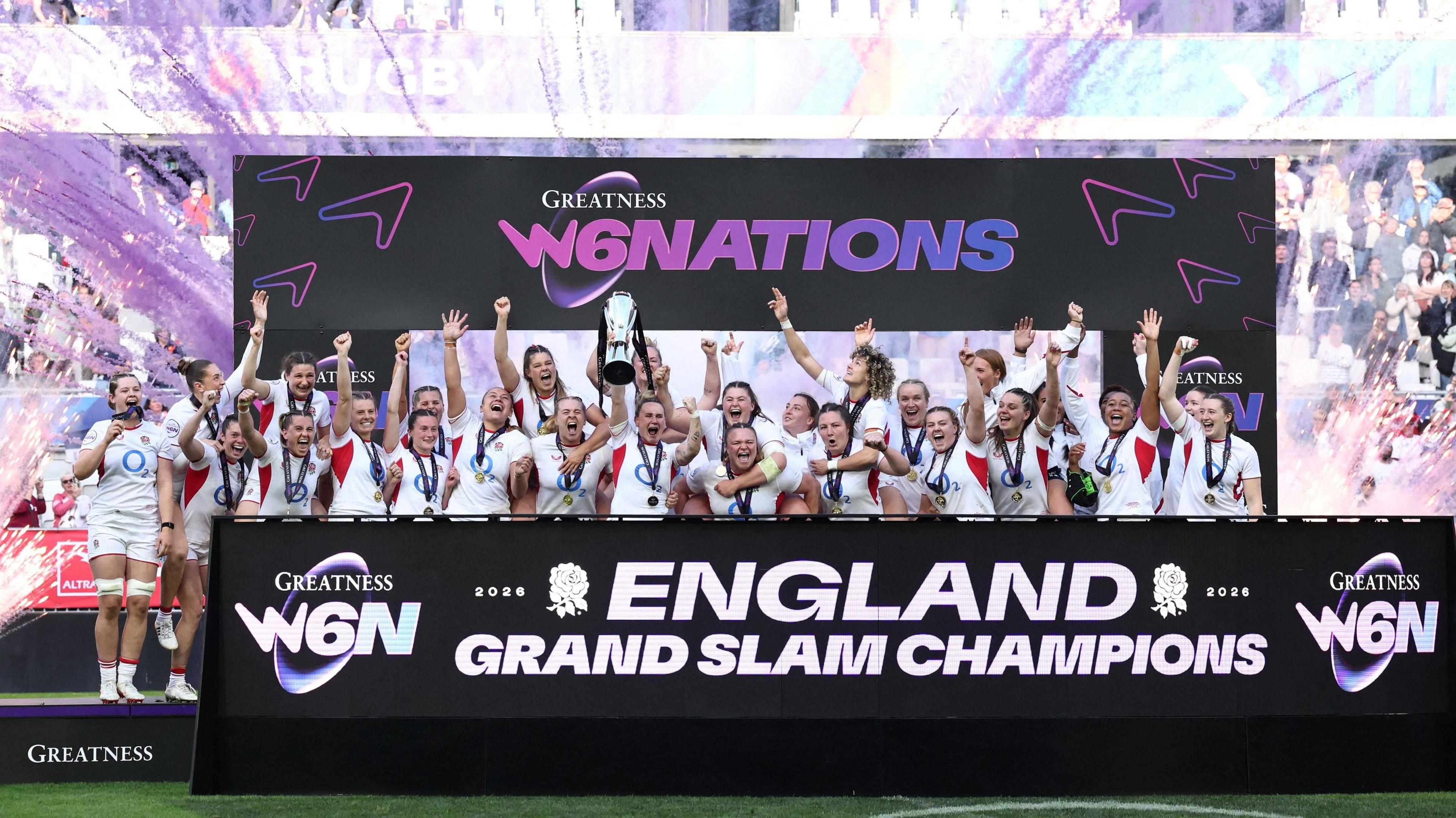 the England women's rugby team celebrating with the six nations trophy with purple flare guns going off behind them