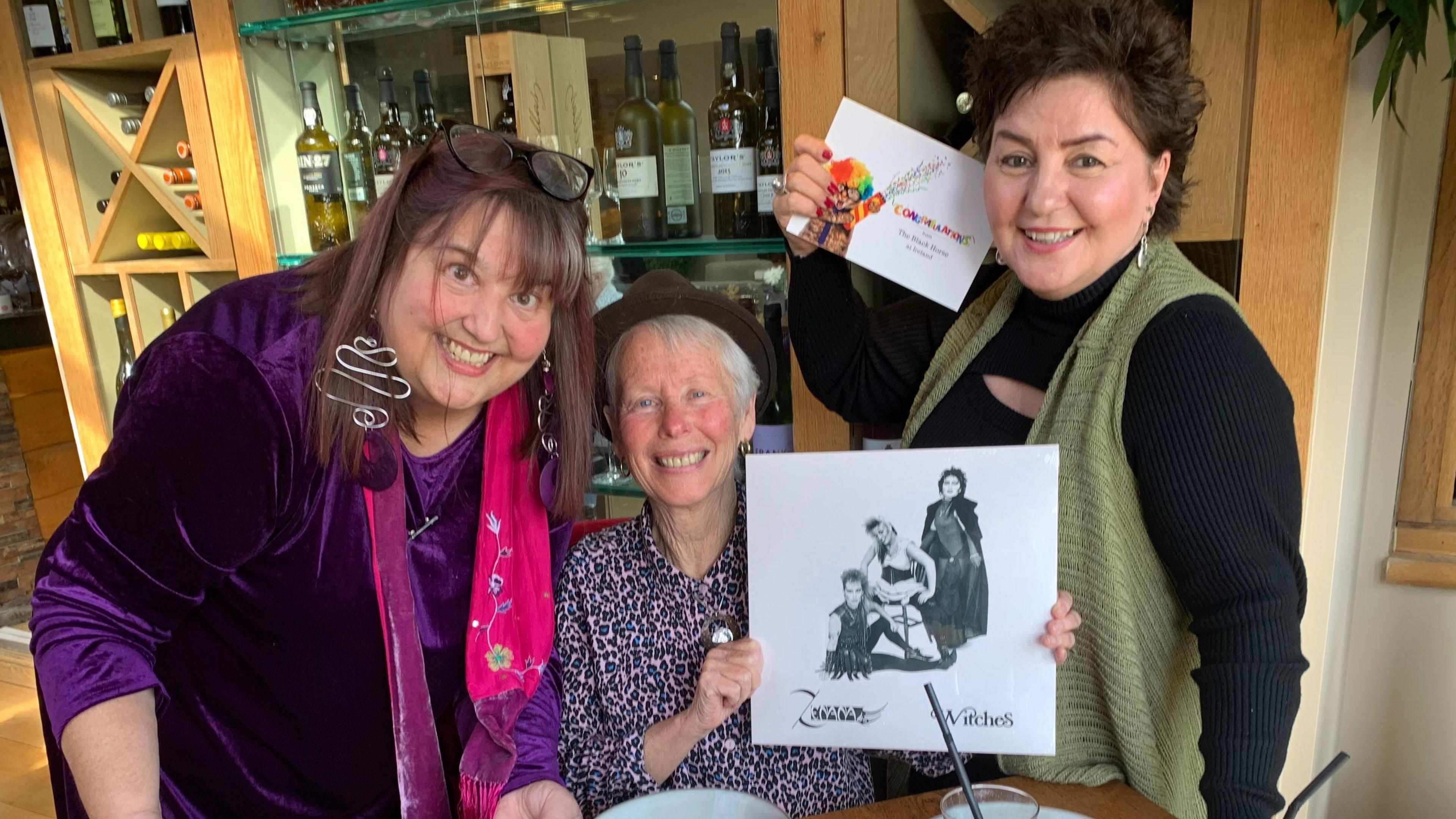 Penny, Anita and Ruth in a room, in front of wine bottles, all smiling and looking at the camera. Anita, in the middle is holding a record of theirs called Witches and Ruth, right is holding a congratulations sign. Anita, is wearing a brown hat,  with a leopard top, Penny a purple top and scarf and Ruth a black top and green jacket. 