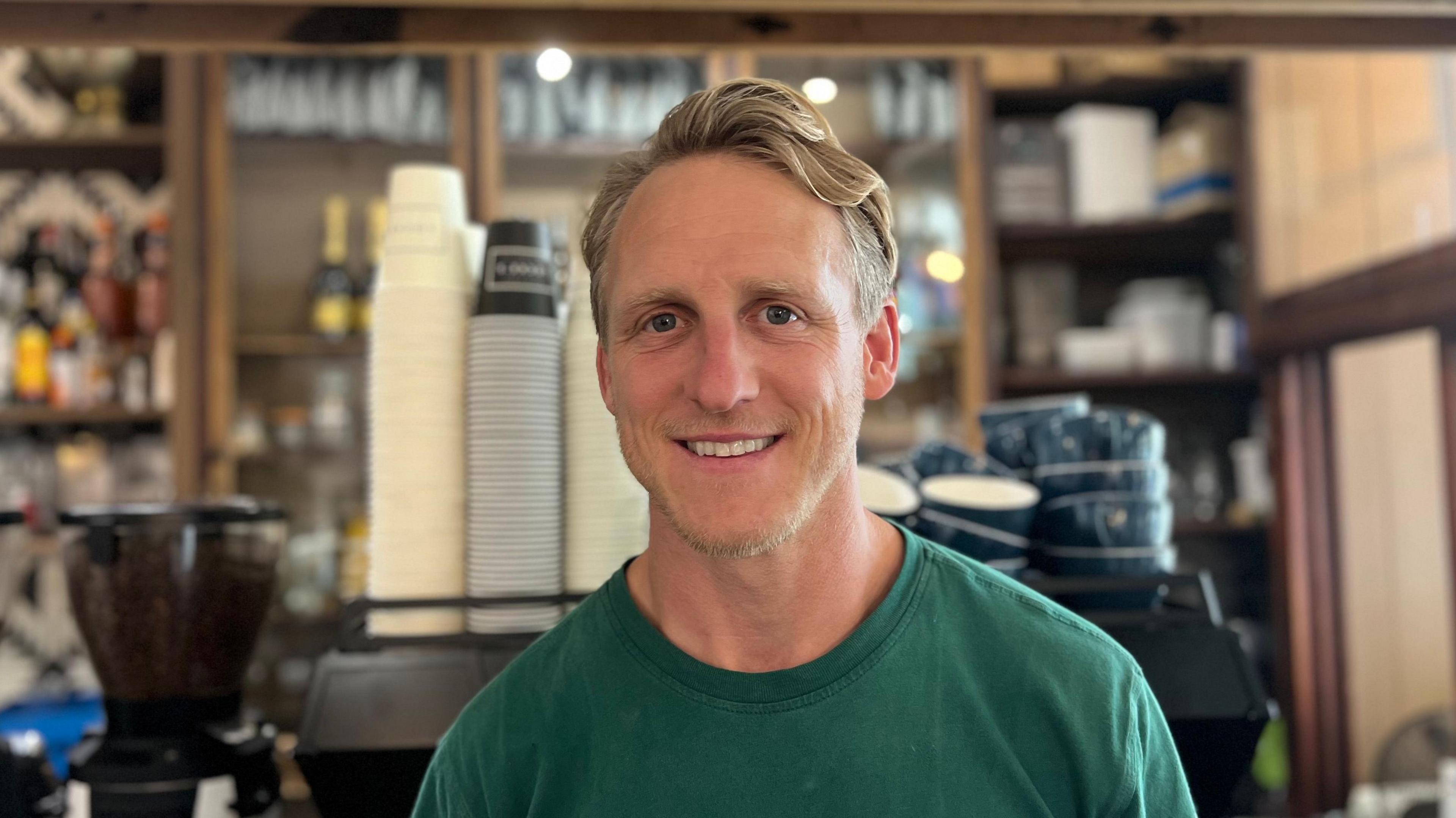 A picture of Drew smiling at the camera. He is wearing a green shirt, has a short beard and blonde hair. He is wearing a green t-shirt. He is stood in front of a coffee machine and a bar.