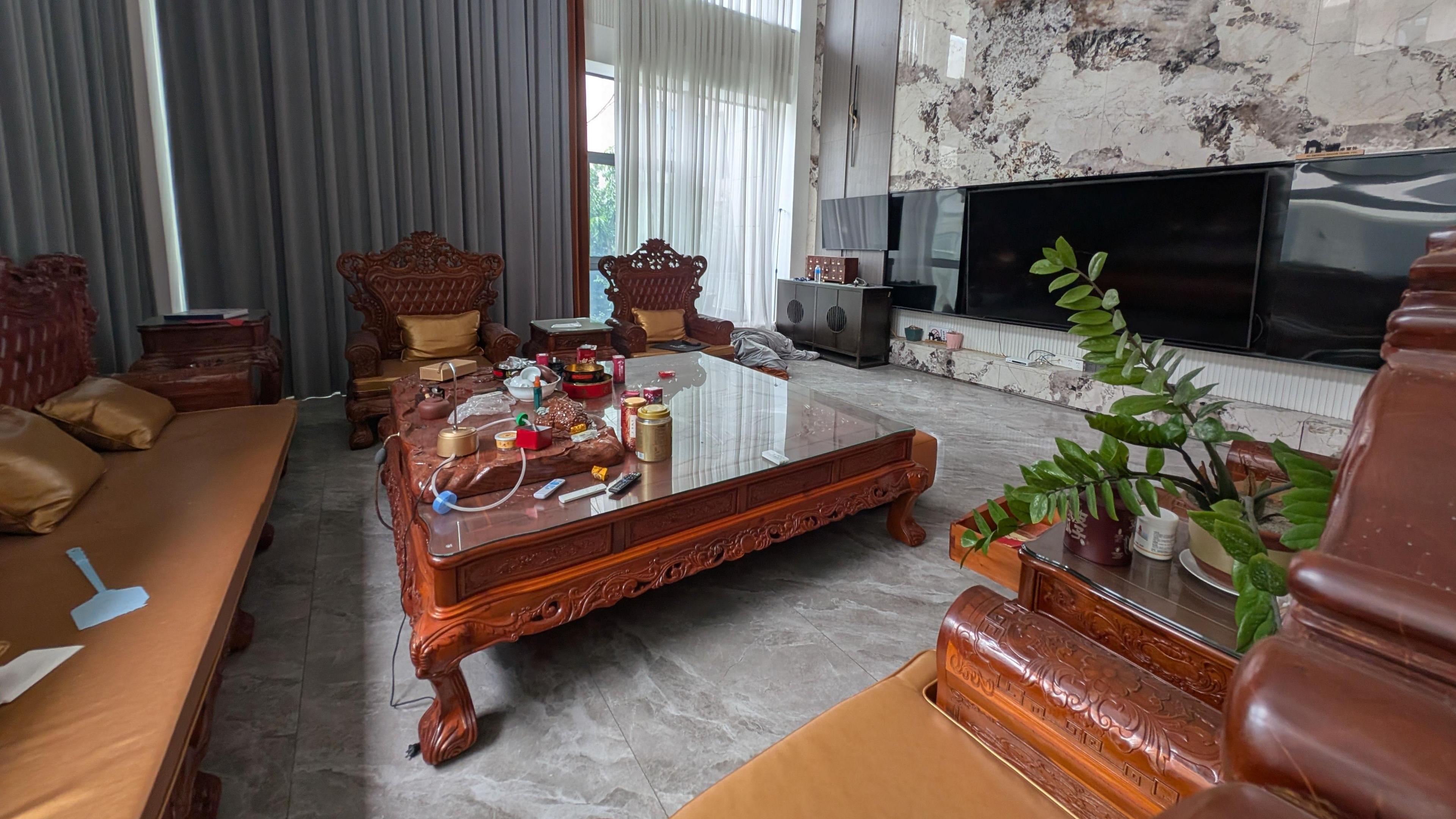 A two-floor-high living room with floor-to-ceiling windows and curtains. The space is clad in marble, and furnished with a coffee table and sofas carved out of wood in a rich red-brown colour. It also features a large flatscreen television.