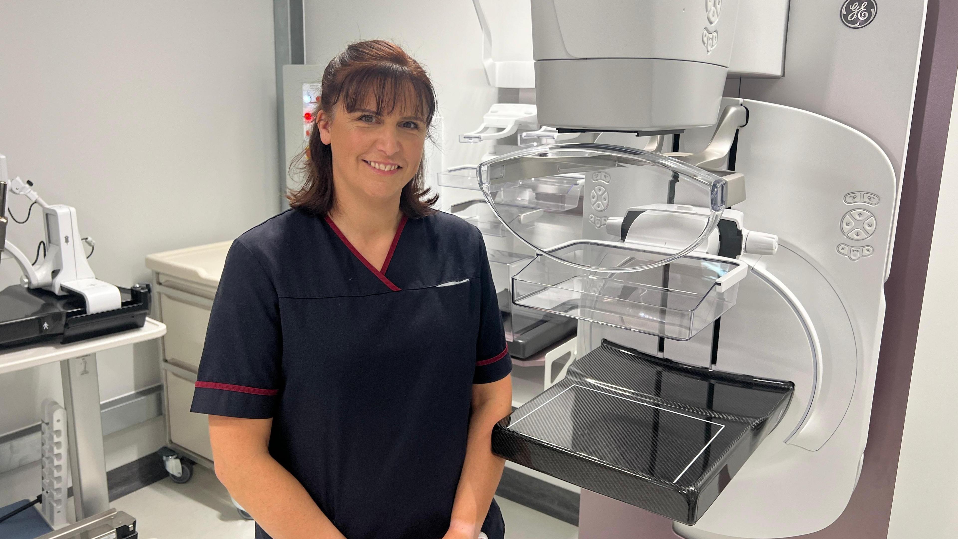 Ms Campbell is standing next to the new equipment at the new breast cancer unit. She is in her scrubs. She has brown hair and a fringe and is smiling.