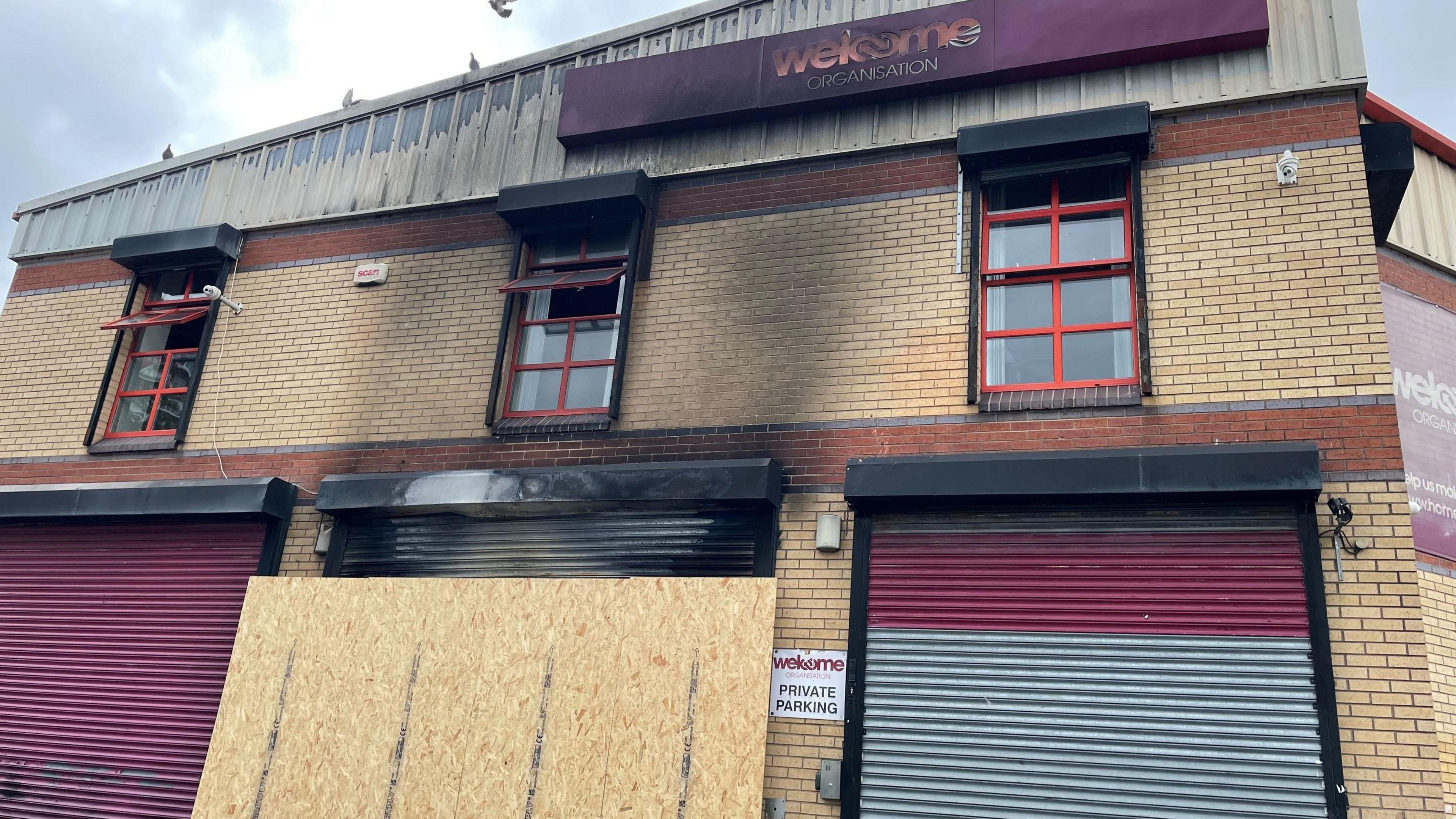 A building with a sign saying 'welcome ORGANISATION' at the top. The outside of the building bears the signs of fire damage, with large black marks on the wall. 